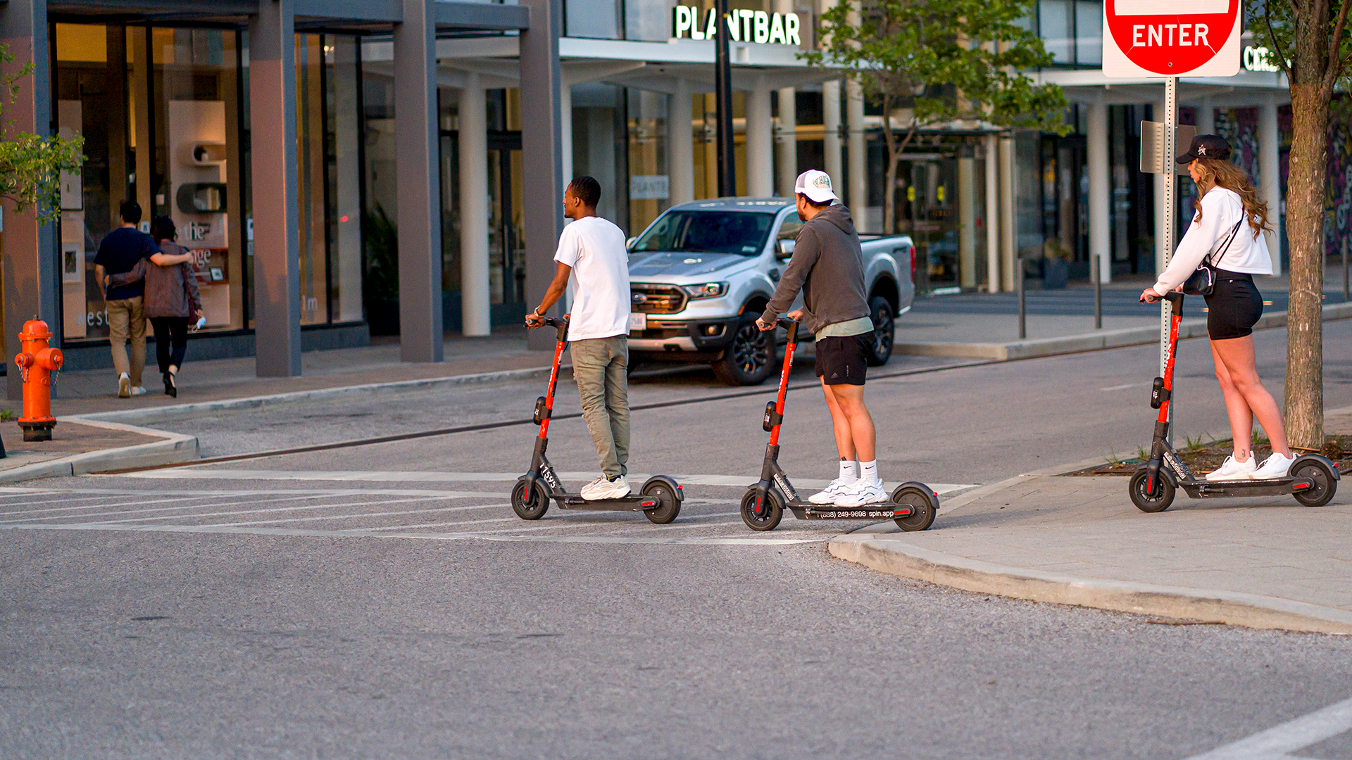 Three people cross the street on e-scooters in Baltimore