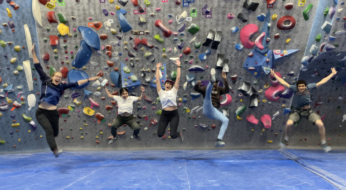 Five people jumping in front of rock climbing wall.
