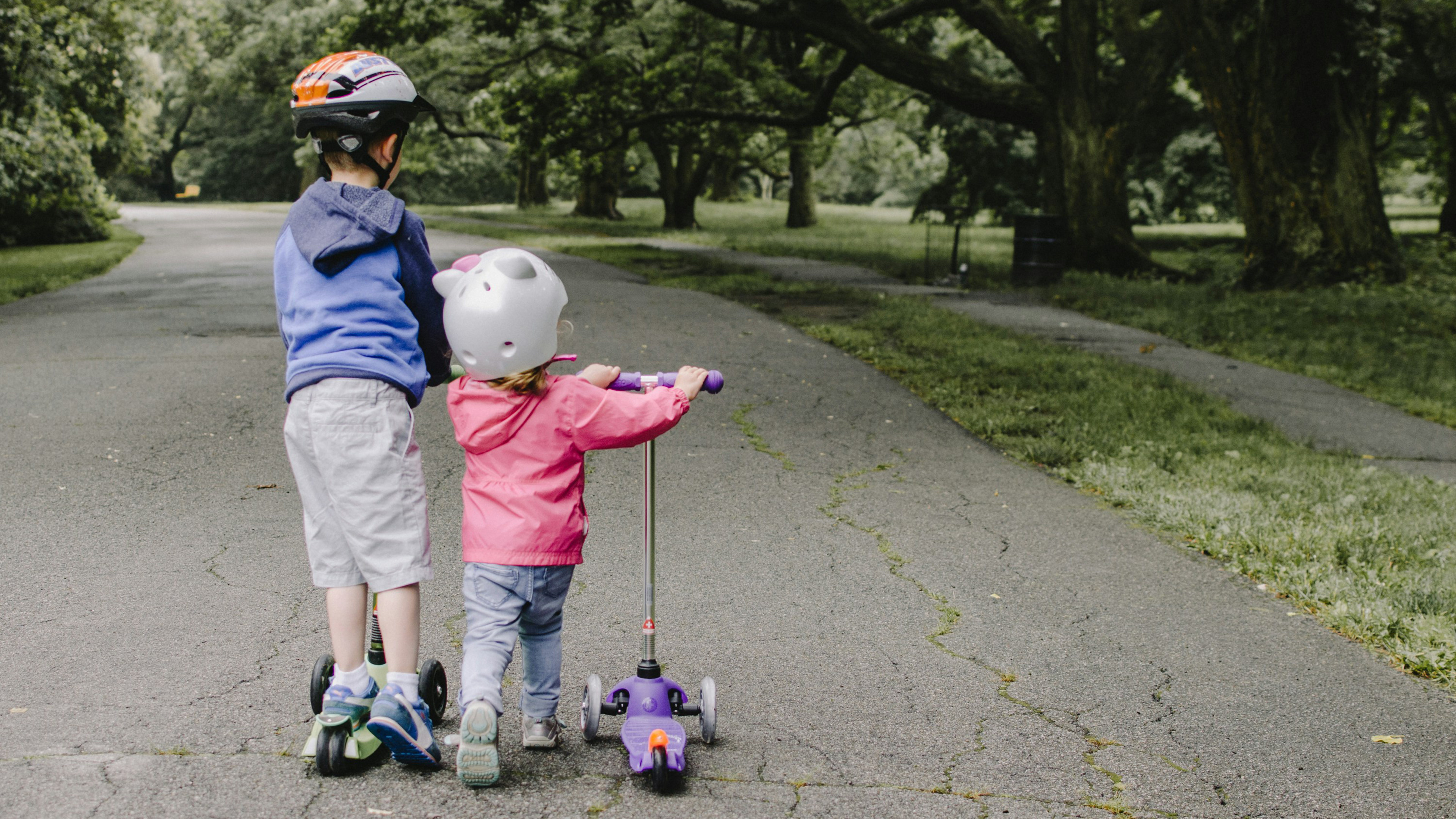 Two children on scooters facing away from the camera as they ride down a road lined with trees.