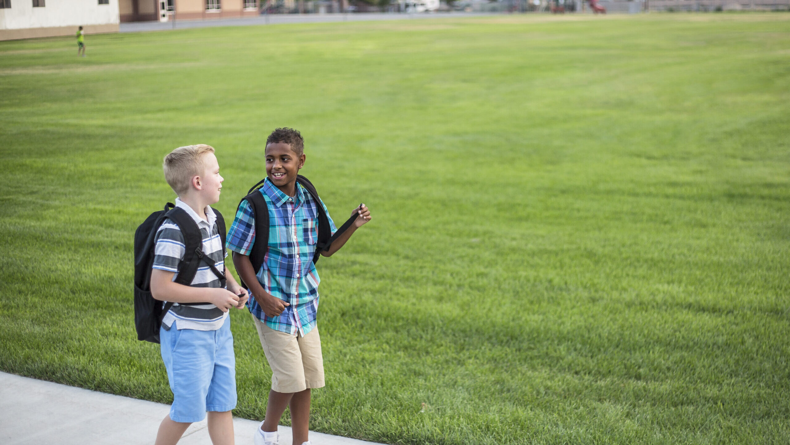 Two diverse school kids walking home together after school and talking together. Back to school photo of diverse school children wearing backpacks in the school yard.