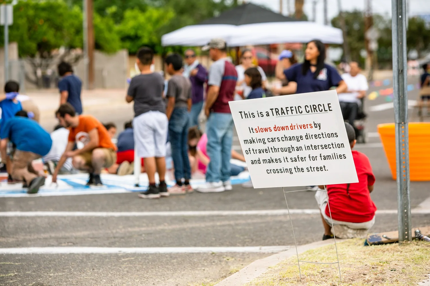 People paint a traffic circle in the middle of an intersection with brightly colored paint. Nearby a sign proclaims: 