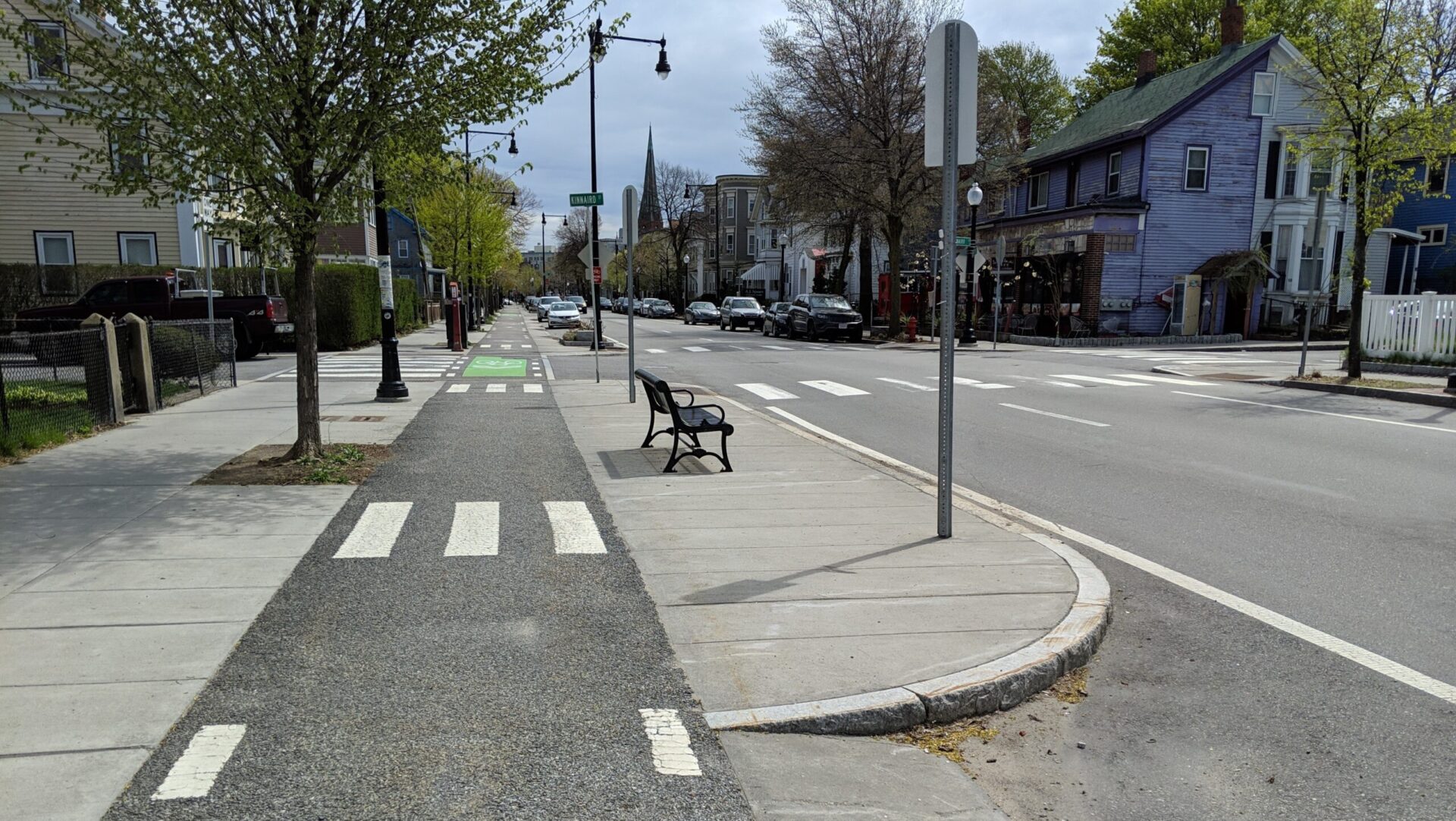 An eye-level photograph of a roadway. The vehicle lanes are separated form a paved bicycle lane by a pedestrian refuge island outfitted with an iron bench.
