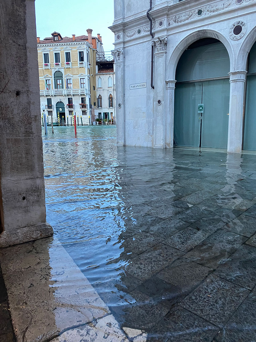 Photograph showing a flooded urban area with water covering stone pavement and reaching building entrances. The scene includes historic architecture with arched doorways and multi-story buildings in the background, highlighting the impact of high water levels on the city environment.