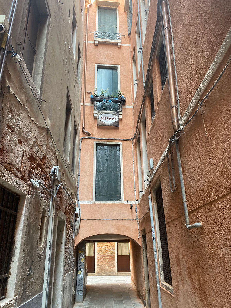 Photograph of a narrow alleyway between tall, weathered buildings with a small archway at the end. The buildings feature peeling paint, exposed brick, pipes along walls, and a window with a flower box above the arch.