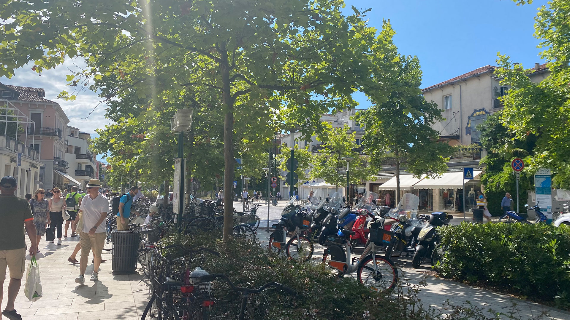 Photograph of a busy urban street scene with pedestrians walking on sidewalks and a row of parked motorcycles and bicycles along the road. The setting features leafy green trees, multi-story buildings with shops and cafes, and bright sunlight casting shadows.