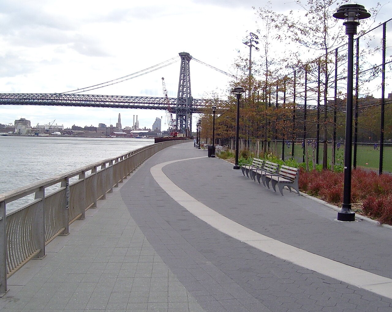 Photograph of a riverside walkway featuring a curved paved path with benches and street lamps along one side and a metal railing along the water. A large suspension bridge spans across the river in the background, with autumn-colored trees and a grassy area adjacent to the path.