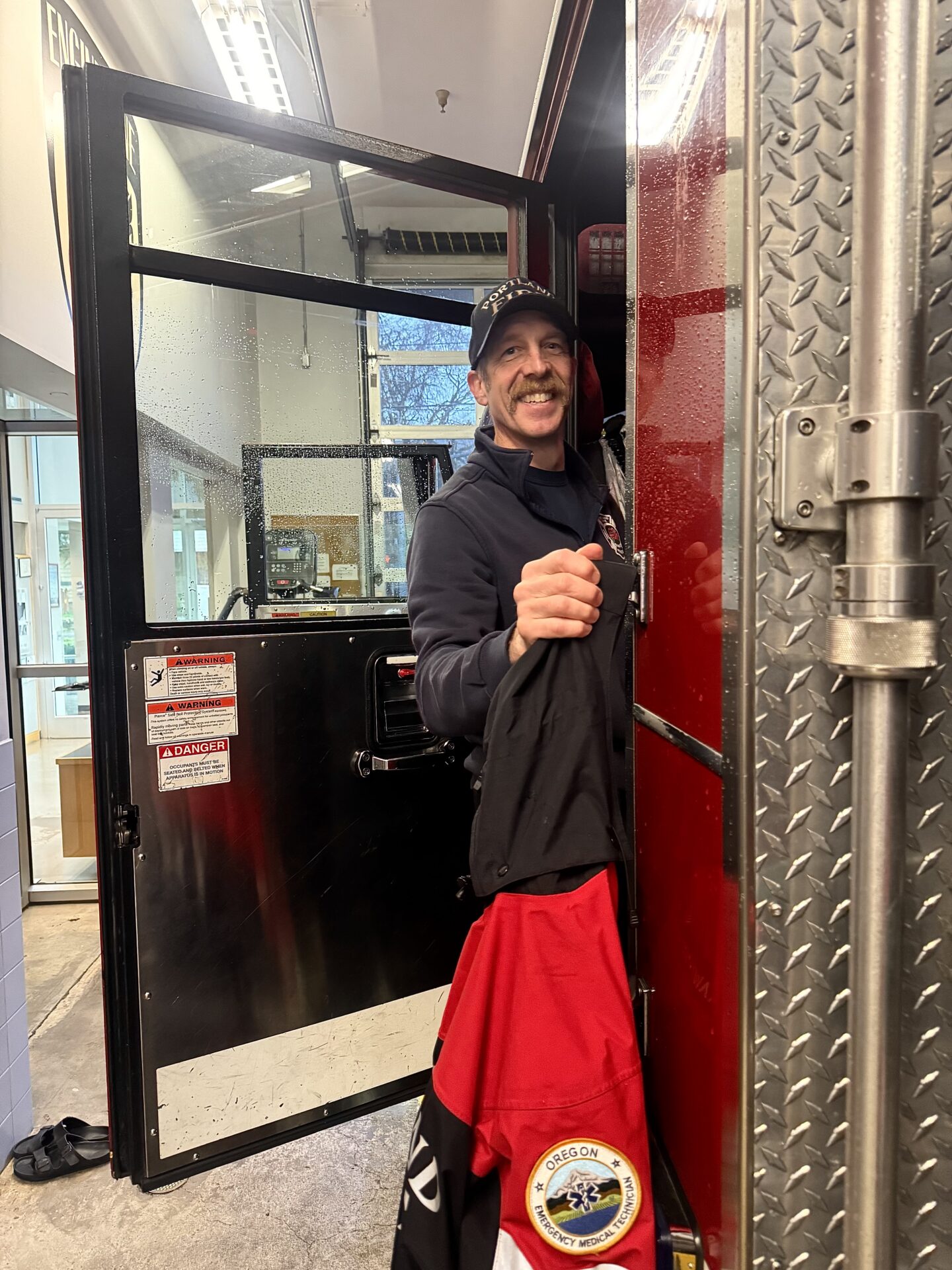 A smiling man with a mustache hangs out of the cab of a fire engine.