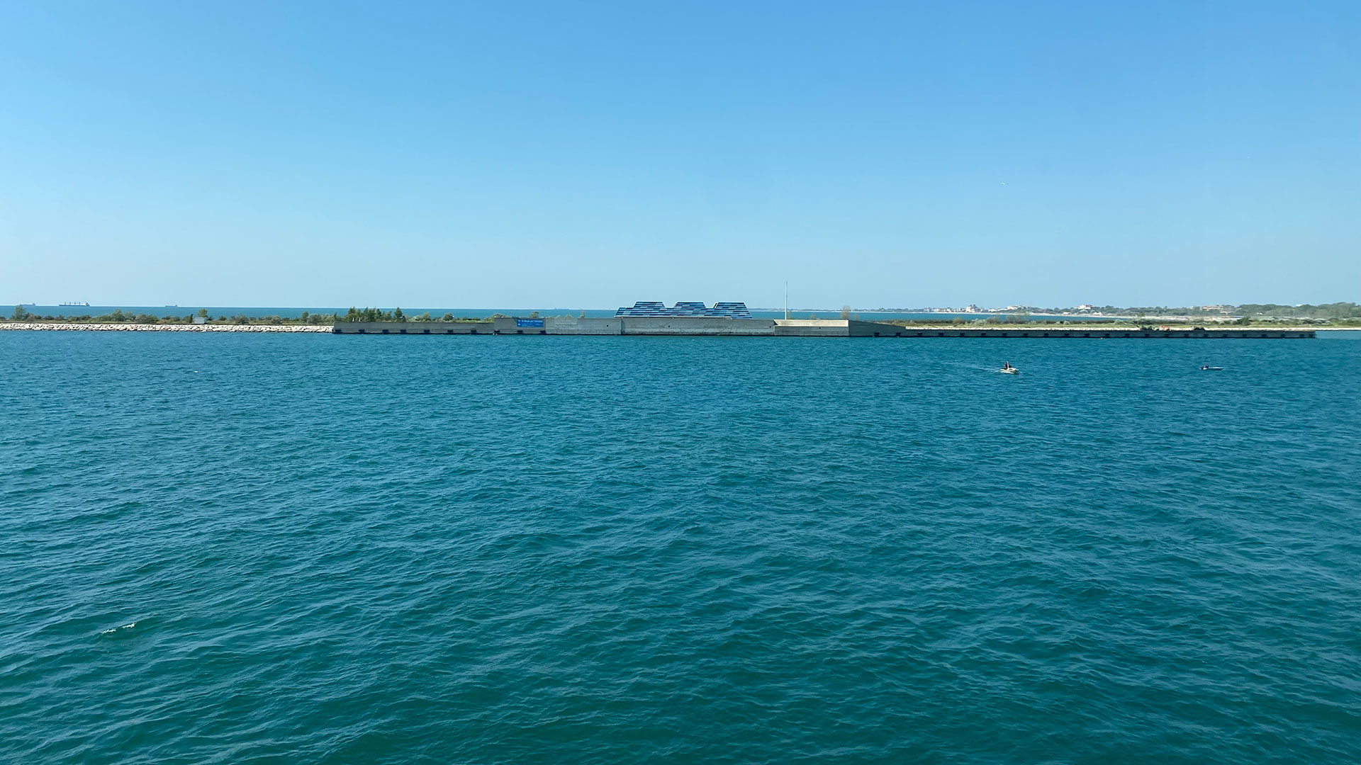 Photograph of a large body of calm blue water with a narrow strip of land and vegetation in the background under a clear blue sky.