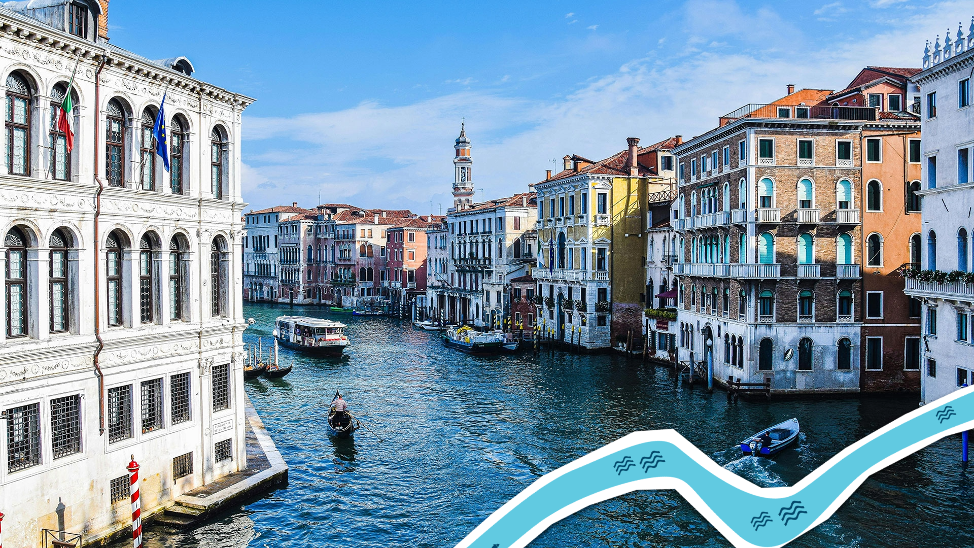 Photograph of a canal in Venice, Italy, showing historic multi-story buildings with colorful facades lining both sides of the waterway. Several boats, including a gondola and motorboats, navigate the canal under a clear blue sky, highlighting the city's iconic waterways and architecture.