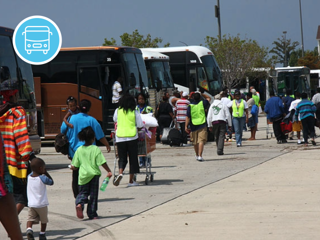 People walk across a parking lot to board buses. Emergency workers wearing neon vests help direct people.