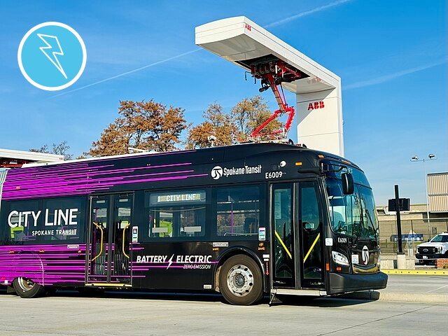 An electric bus is parked under an overhead charger, which connects to its roof to supply power.
