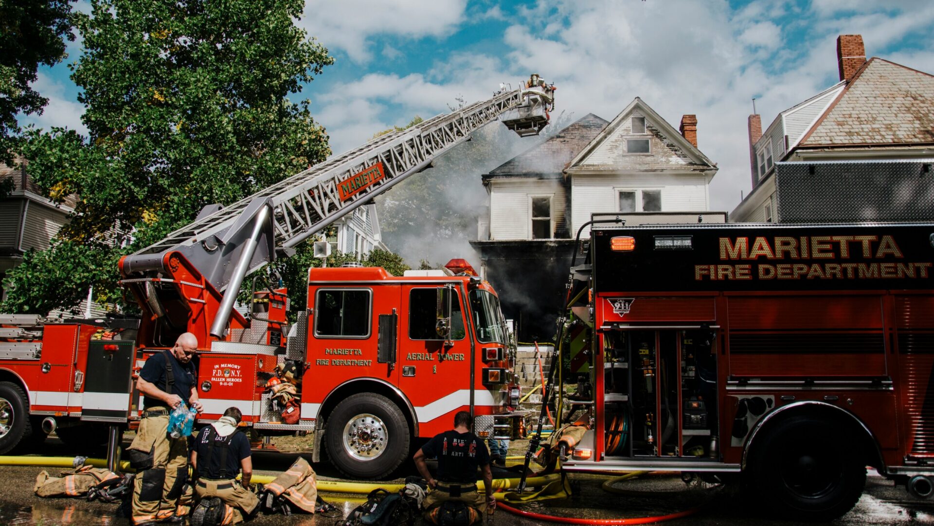 A crew works to put out a house fire. Firefighters kneel next to a red fire truck with its ladder extended while smoke billows out from the house behind.