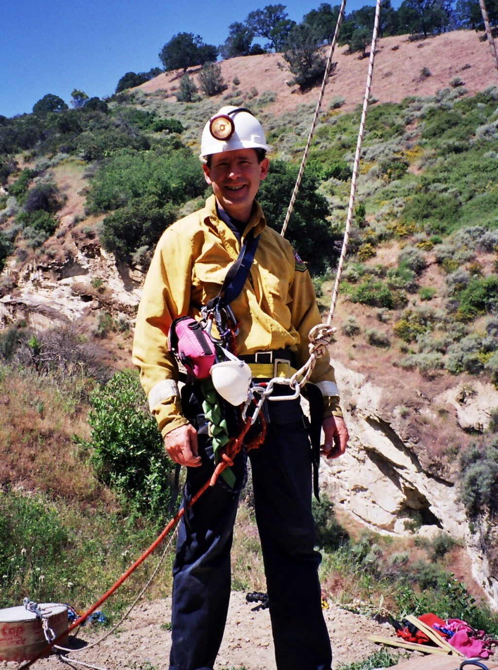 A smiling man in hard hat, safety jacket, and climbing rigging stands in front of California hills.