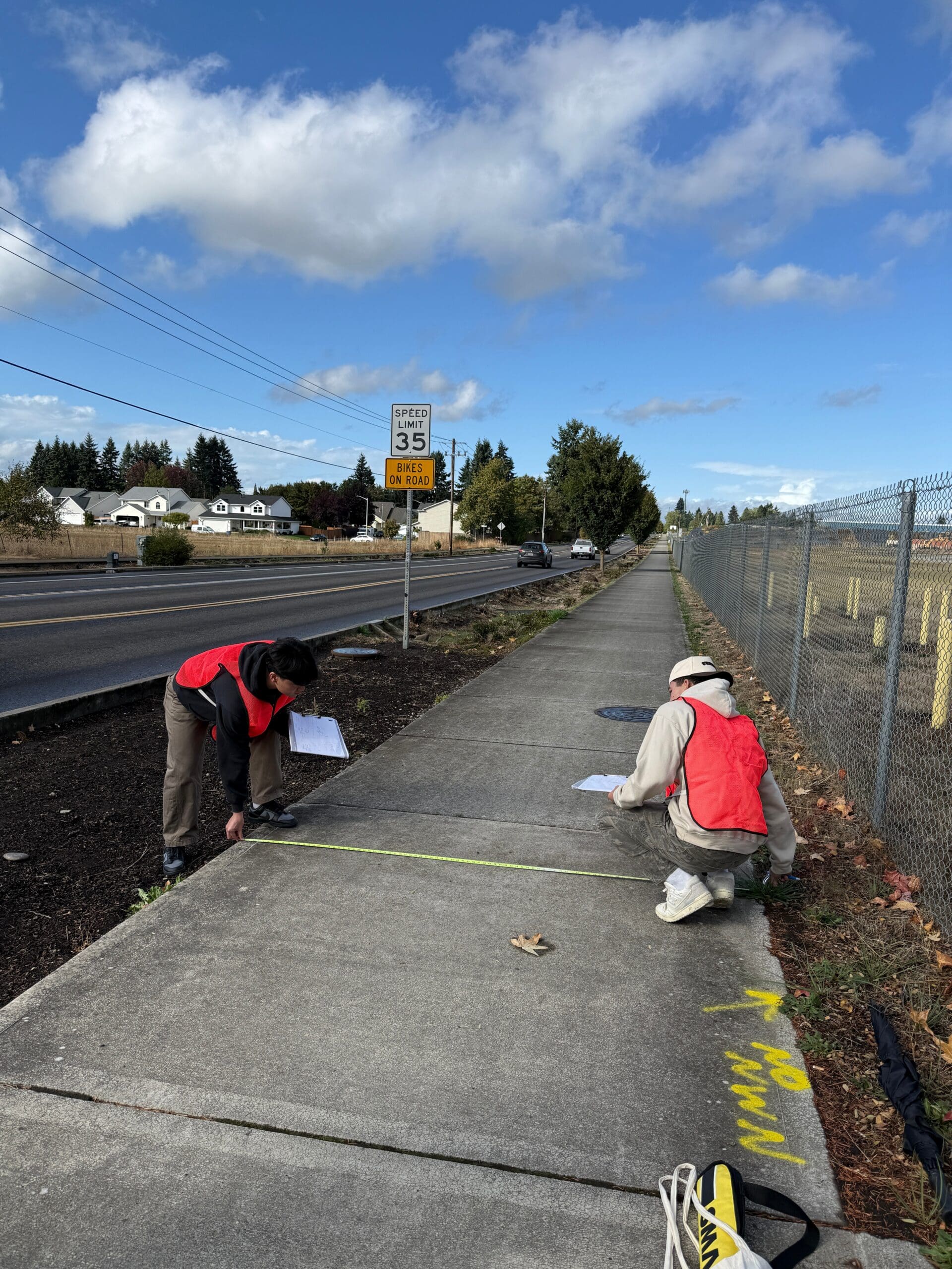 Photograph showing two people wearing orange safety vests measuring and marking a sidewalk next to a road with a 35 mph speed limit sign.