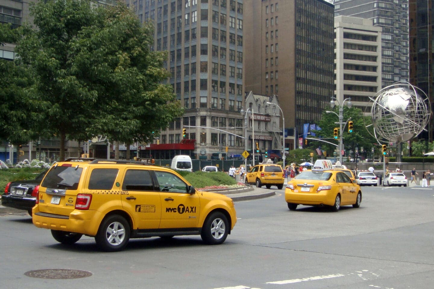 Photograph of a busy urban street scene featuring two yellow taxis driving near a large spherical metal sculpture with office buildings, trees, pedestrians, and traffic signals.