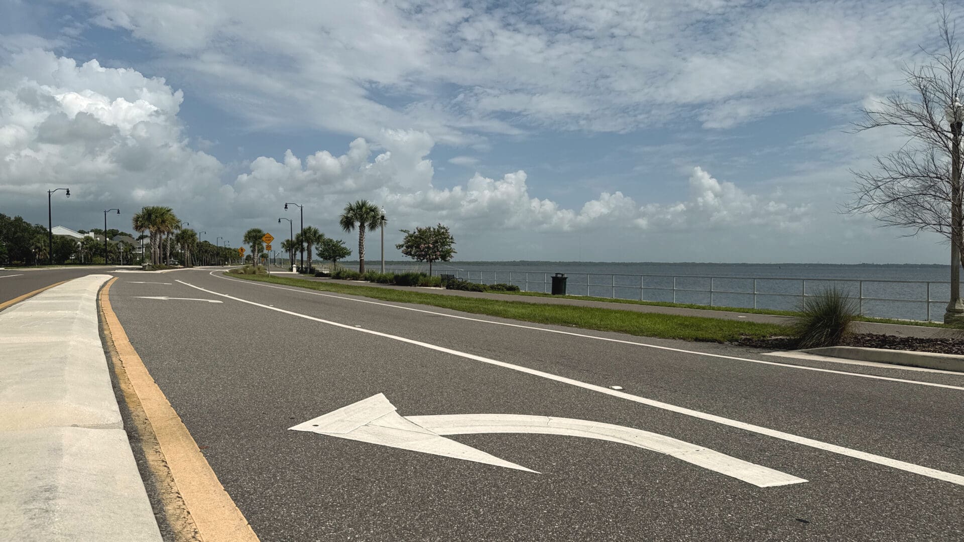 Photograph of a curved road with a large white left-turn arrow painted on asphalt, bordered by a sidewalk and a waterfront with a metal railing.