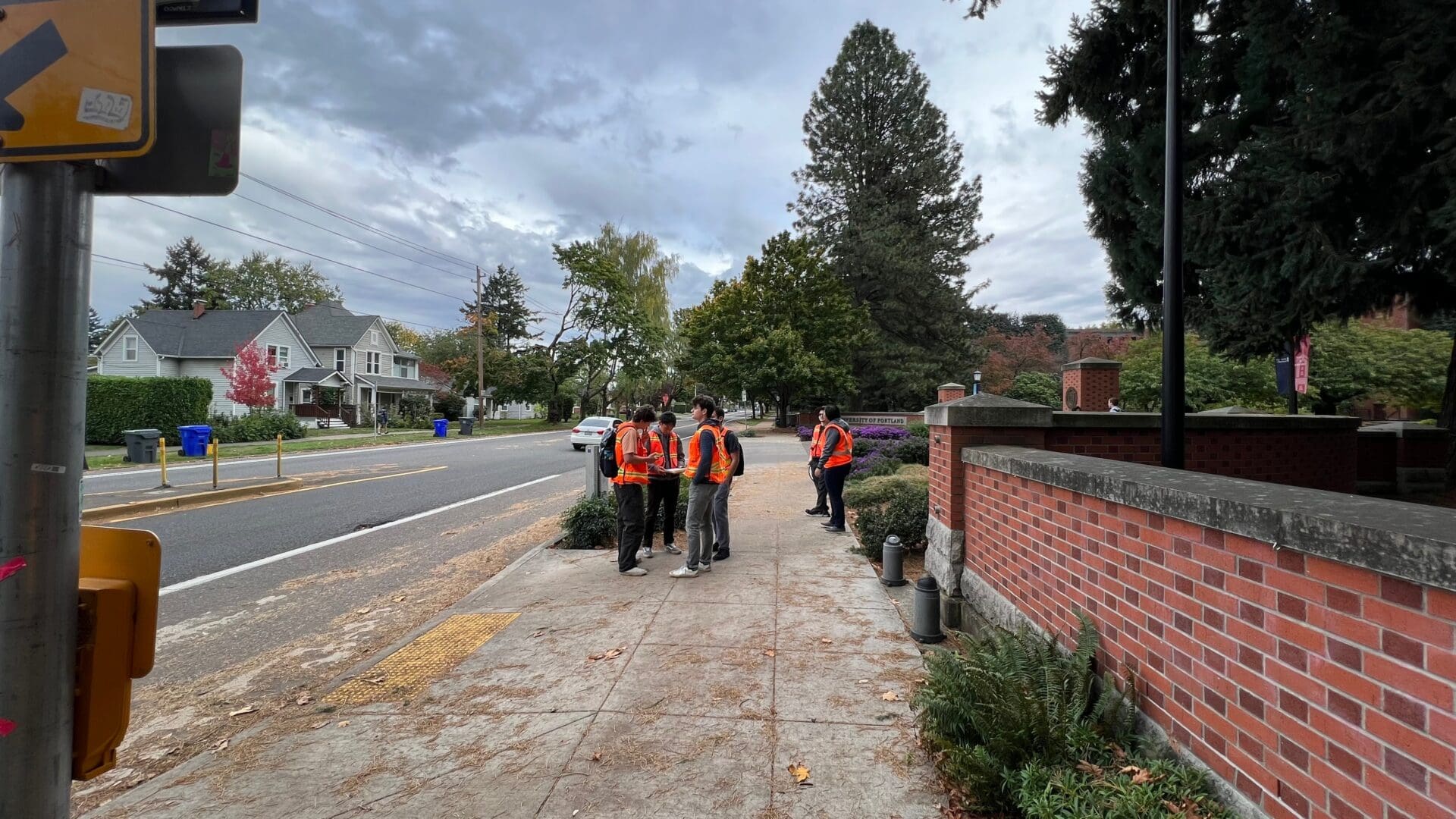 Photograph showing a group of students wearing orange safety vests standing on a sidewalk next to a red brick wall and a street lined with houses and trees.