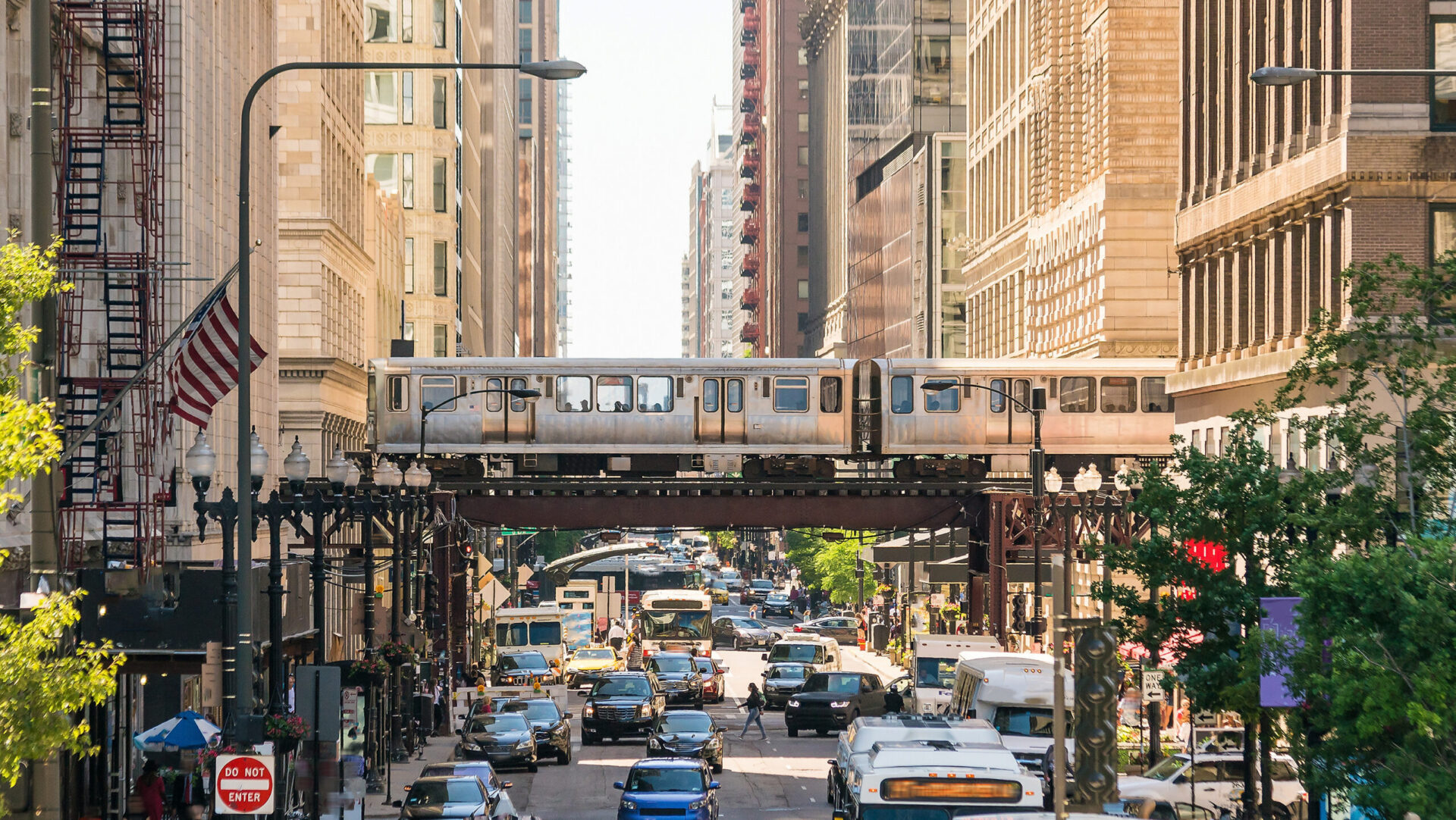 A train passes on an elevated rail line over a busy street. Cars, buses, and pedestrians travel below, amidst tall buildings.