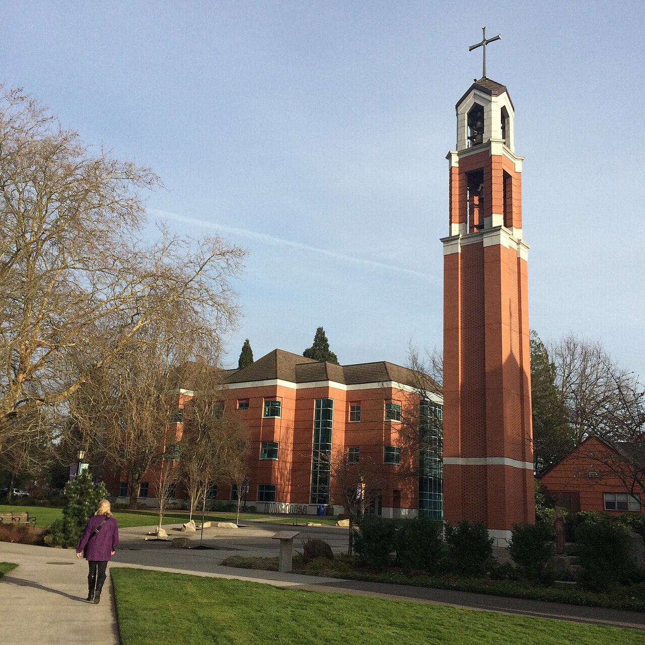 Photograph of a tall brick bell tower with a cross on top, situated near a modern brick building with large windows and surrounded by trees and greenery. A person wearing a purple jacket walks on a paved path leading toward the building, under a clear blue sky.