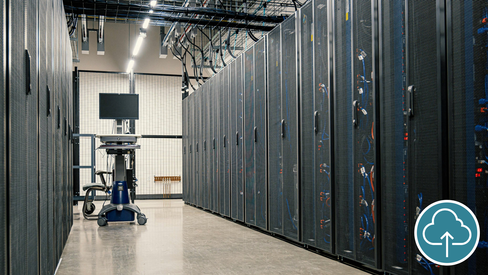 Data center aisle featuring rows of black server racks with mesh doors on both sides. A mobile workstation with a monitor and keyboard is positioned at the end of the aisle.
