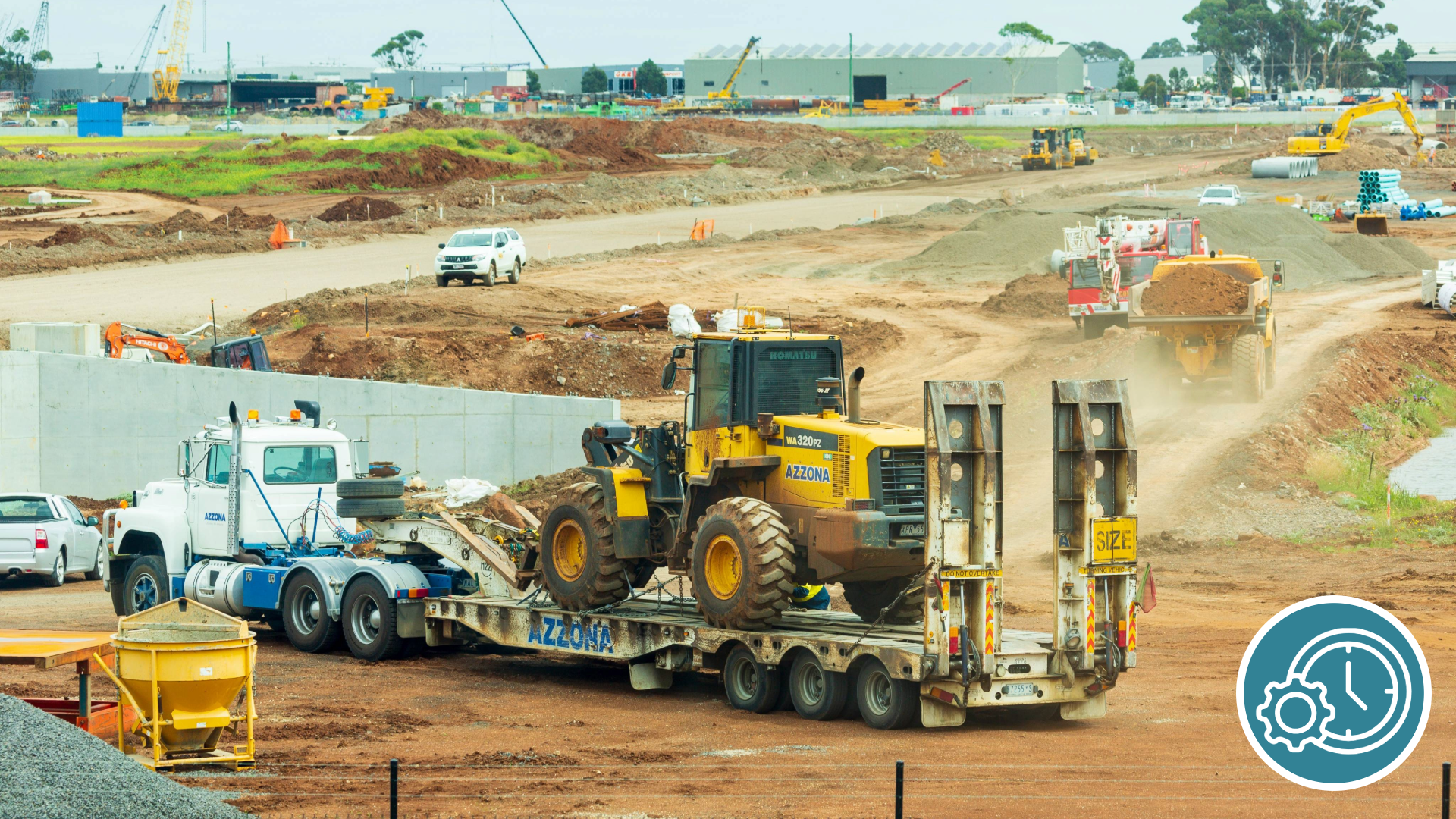 Construction site showing heavy machinery being transported on a flatbed trailer attached to a white truck. There are dirt roads, construction equipment, and cranes in the background.