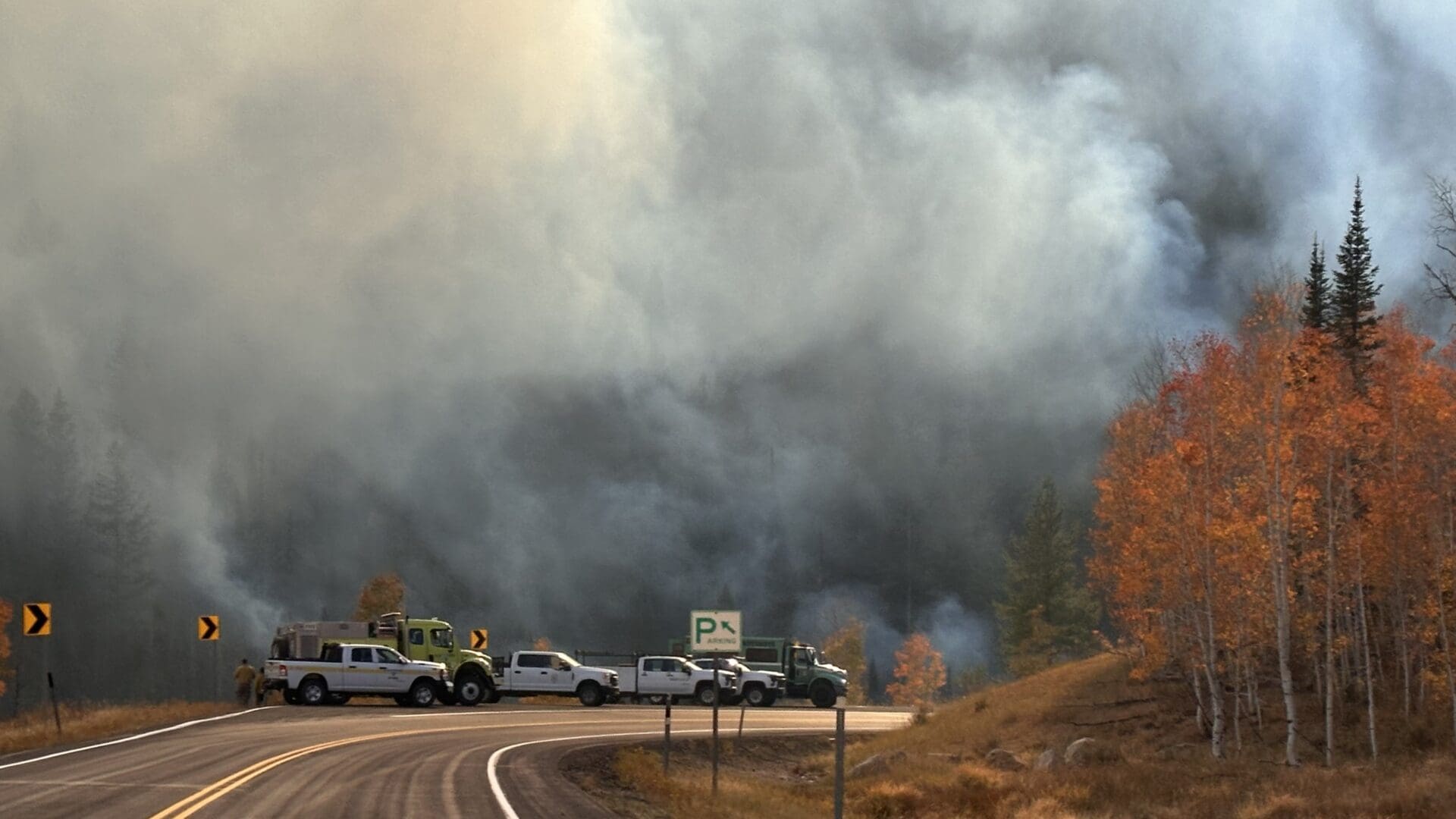 Photograph showing a wildfire with thick smoke billowing in a forested area during autumn, with trees displaying orange and yellow leaves. Several vehicles, including emergency response trucks, are parked along a road.