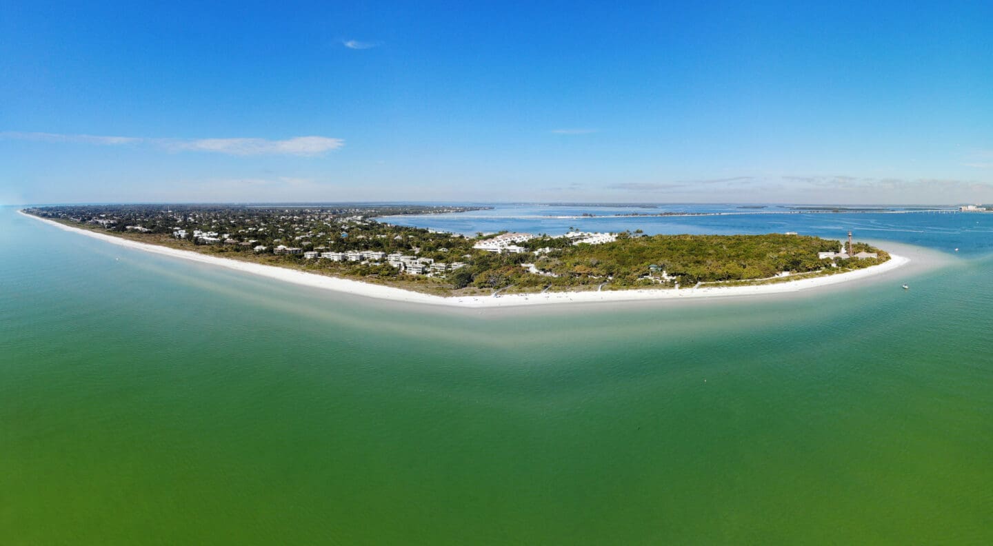 Aerial photograph of a narrow island surrounded by greenish-blue water under a clear blue sky. The island features dense vegetation, scattered buildings, and sandy beaches along its shoreline.