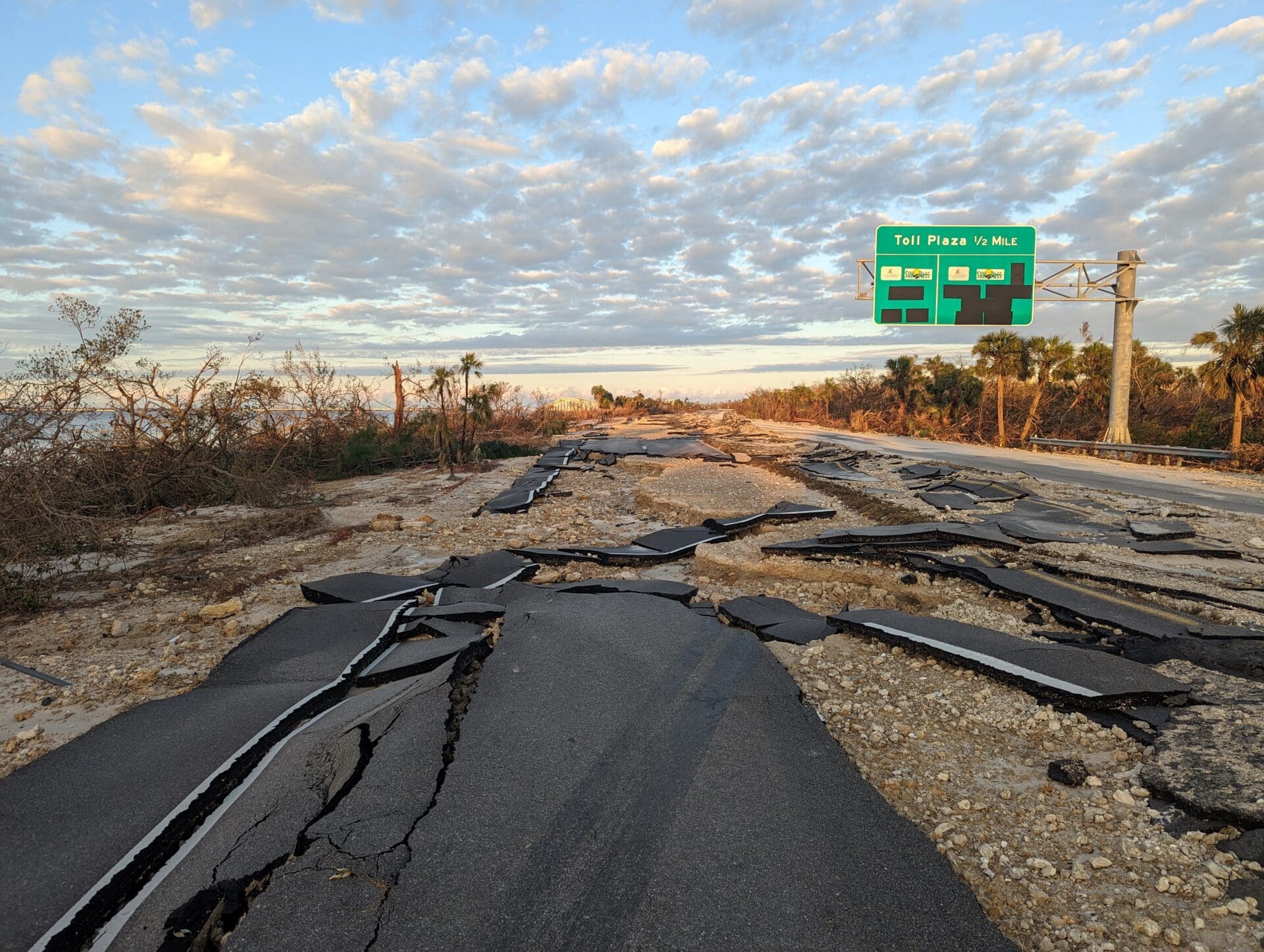 Photograph of a severely damaged road with cracked and displaced asphalt under a partly cloudy sky.