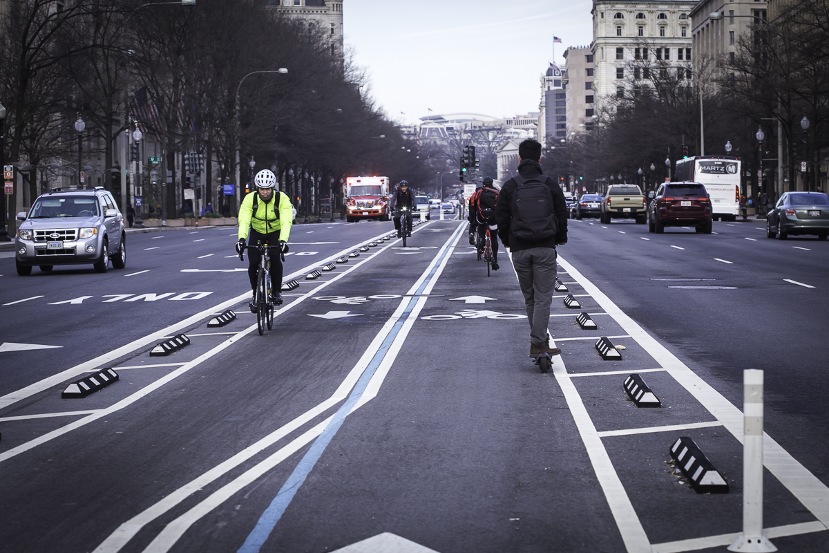 Photograph of an urban street showing a separated bike lane with cyclists and an e-scooter using the designated lanes in the middle of the road with vehicle lanes on either side. The scene includes traffic barriers, road markings in white and blue, and surrounding buildings.