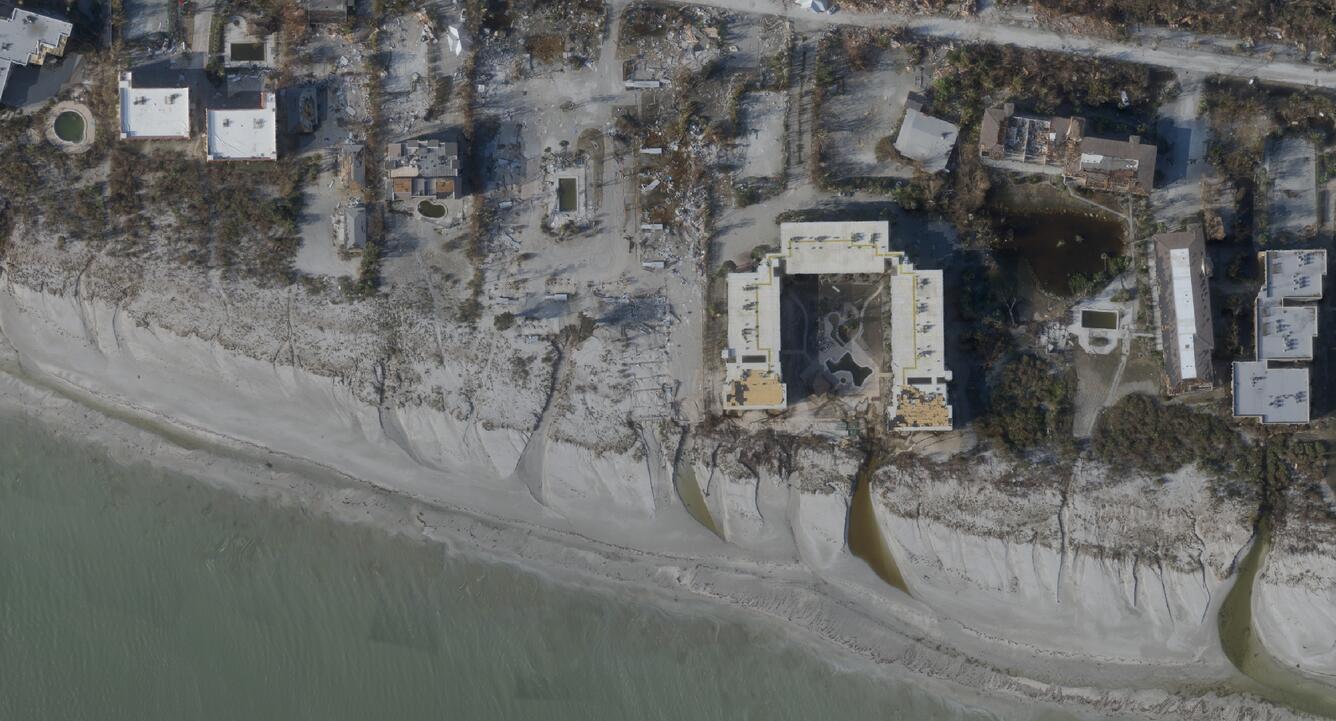 Aerial photograph showing coastal erosion affecting buildings along a shoreline. Sand has been washed away, exposing foundations and creating channels where water flows from the land to the sea.