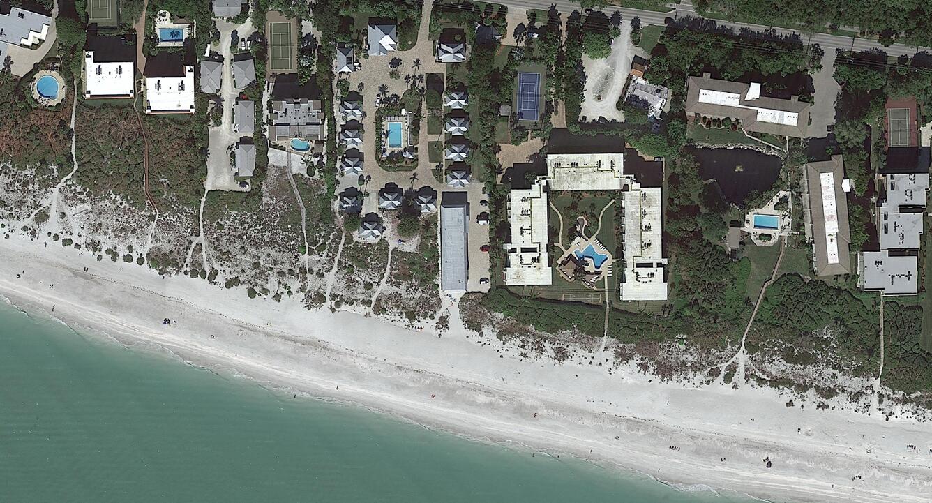 Aerial photograph showing a coastal area with a sandy beach along the bottom and residential buildings, including houses and apartment complexes, near the top. Several properties feature swimming pools, and pathways connect the beach to the residences.