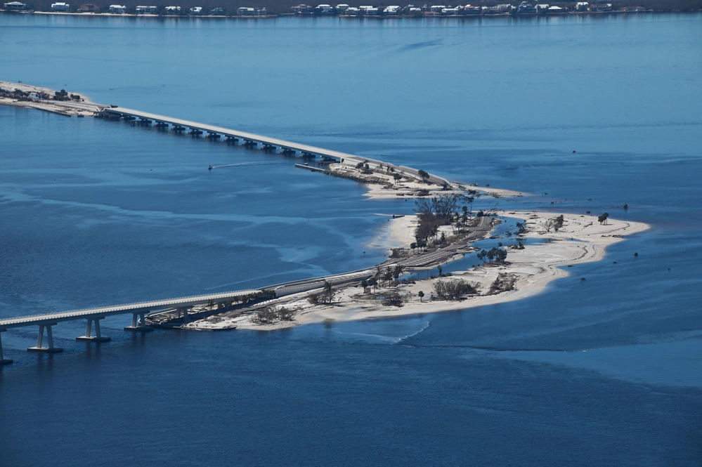 Aerial photograph showing a long bridge extending over a body of water with a partially submerged sandbar or small island beneath it. The water levels cover parts of the land.