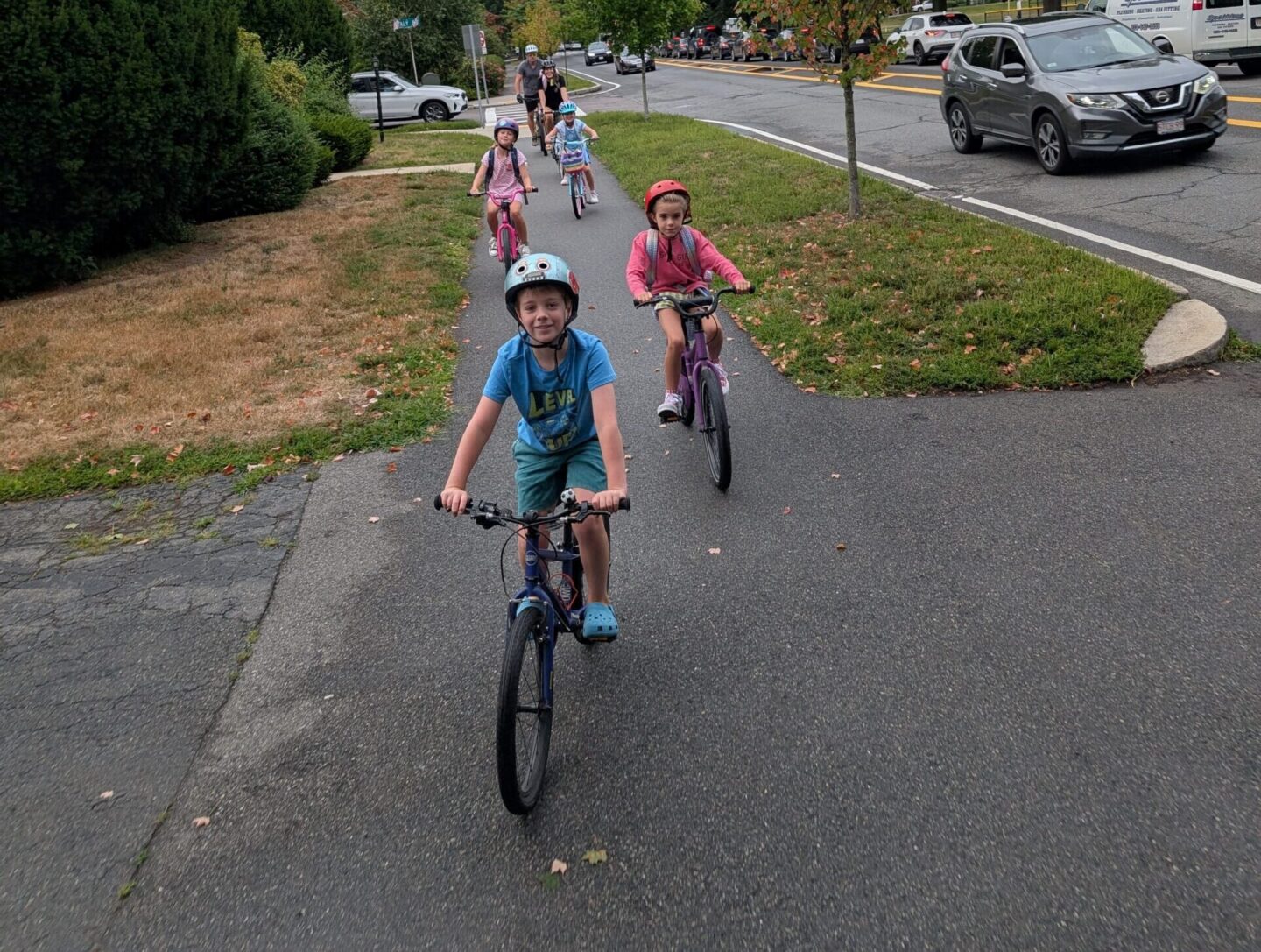 A small group of children ride bikes on a paved path next to a road with cars.