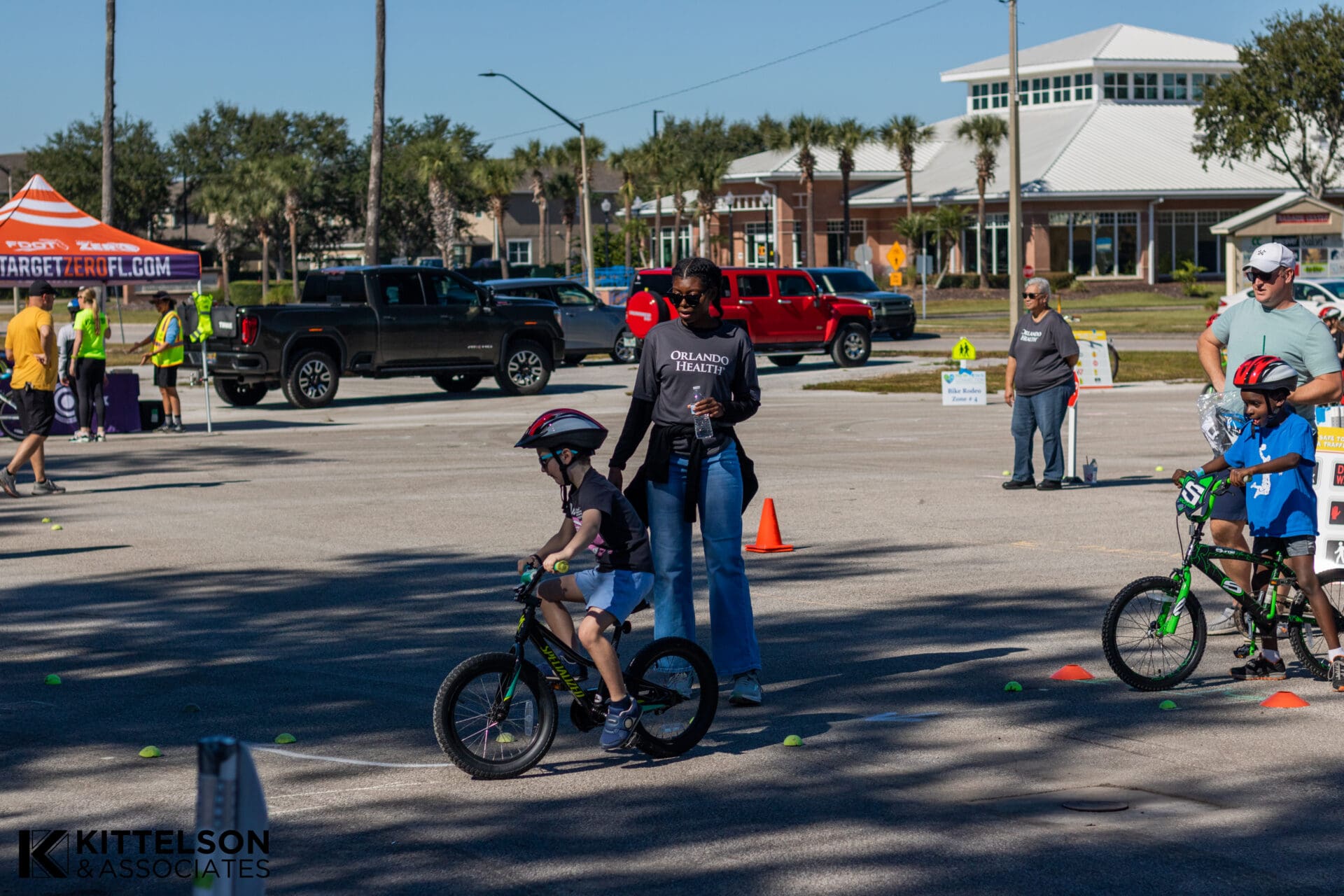 Children ride bikes through paths designated by cones and tennis balls. Adults stand by to supervise.
