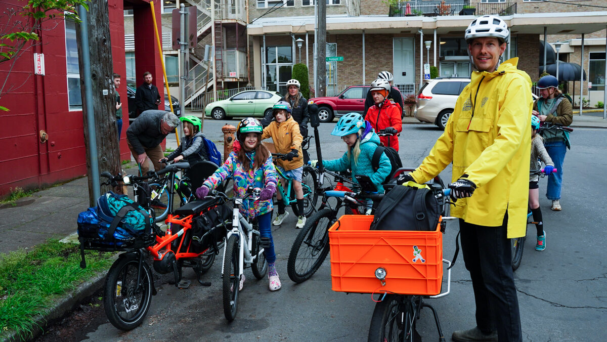 On a neighborhood street, an adult with a bike stands next to a group of children getting on their bikes.