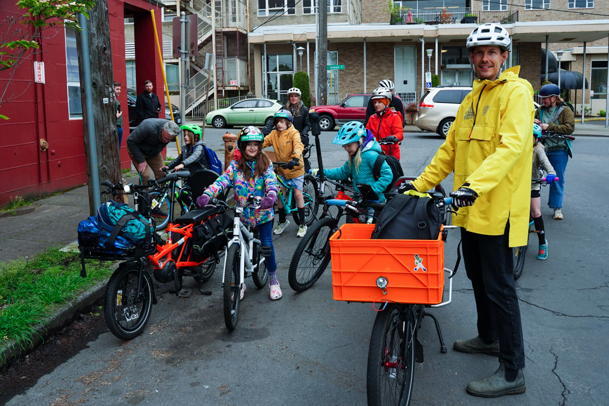 On a neighborhood street, an adult with a bike stands next to a group of children getting on their bikes.