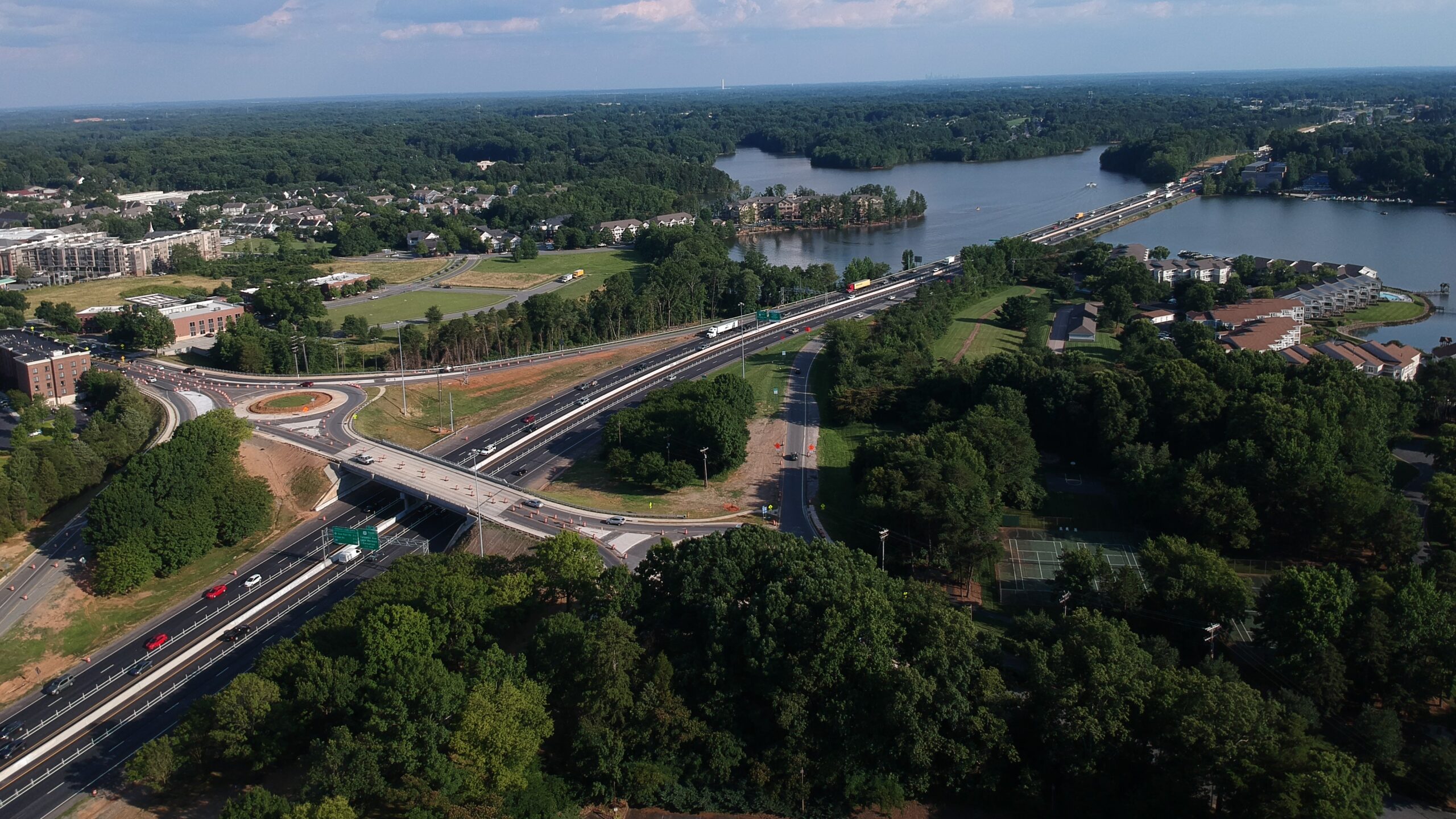 Aerial photograph of a highway bridge crossing over a river surrounded by dense trees and residential areas. The highway includes a roundabout on one side of the bridge and multiple vehicles on the road.
