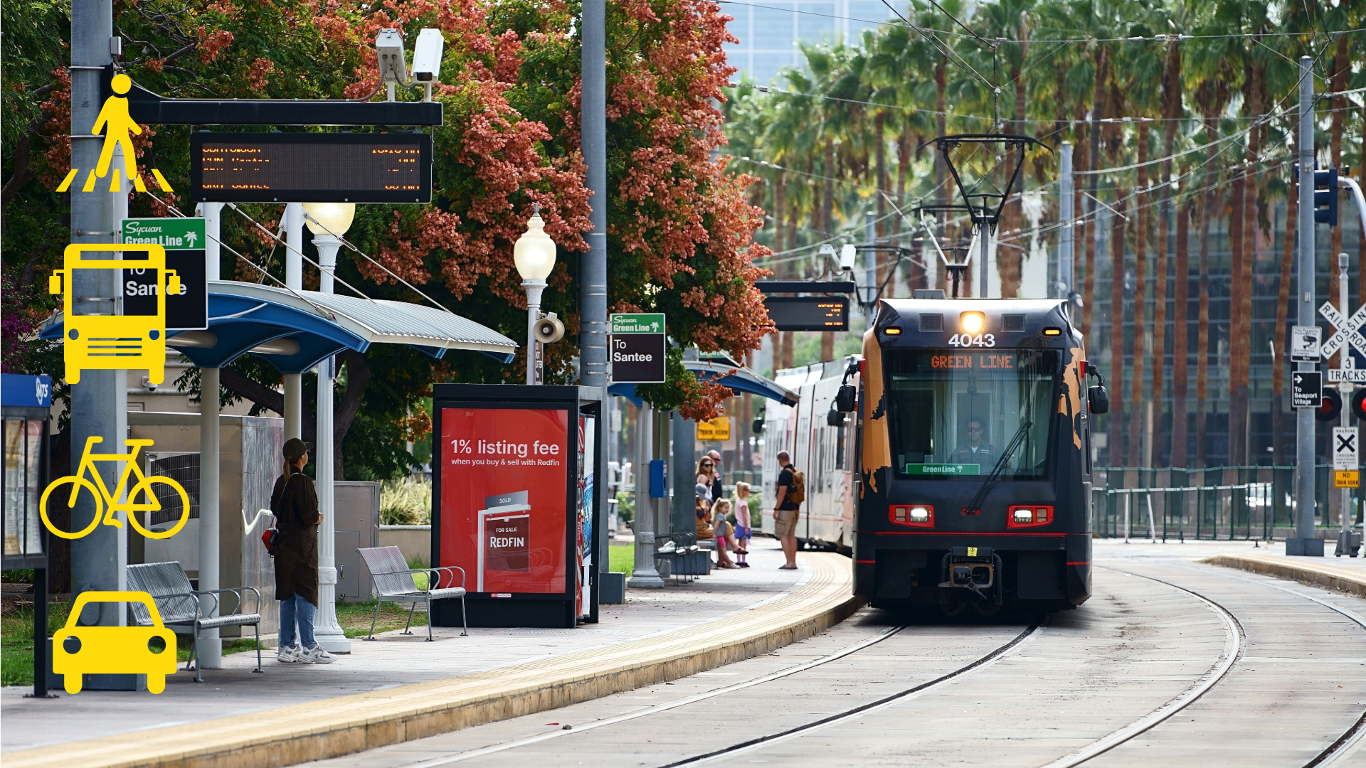 Photograph of a streetcar approaching a station platform with passengers waiting under blue canopies and surrounded by trees and urban buildings.