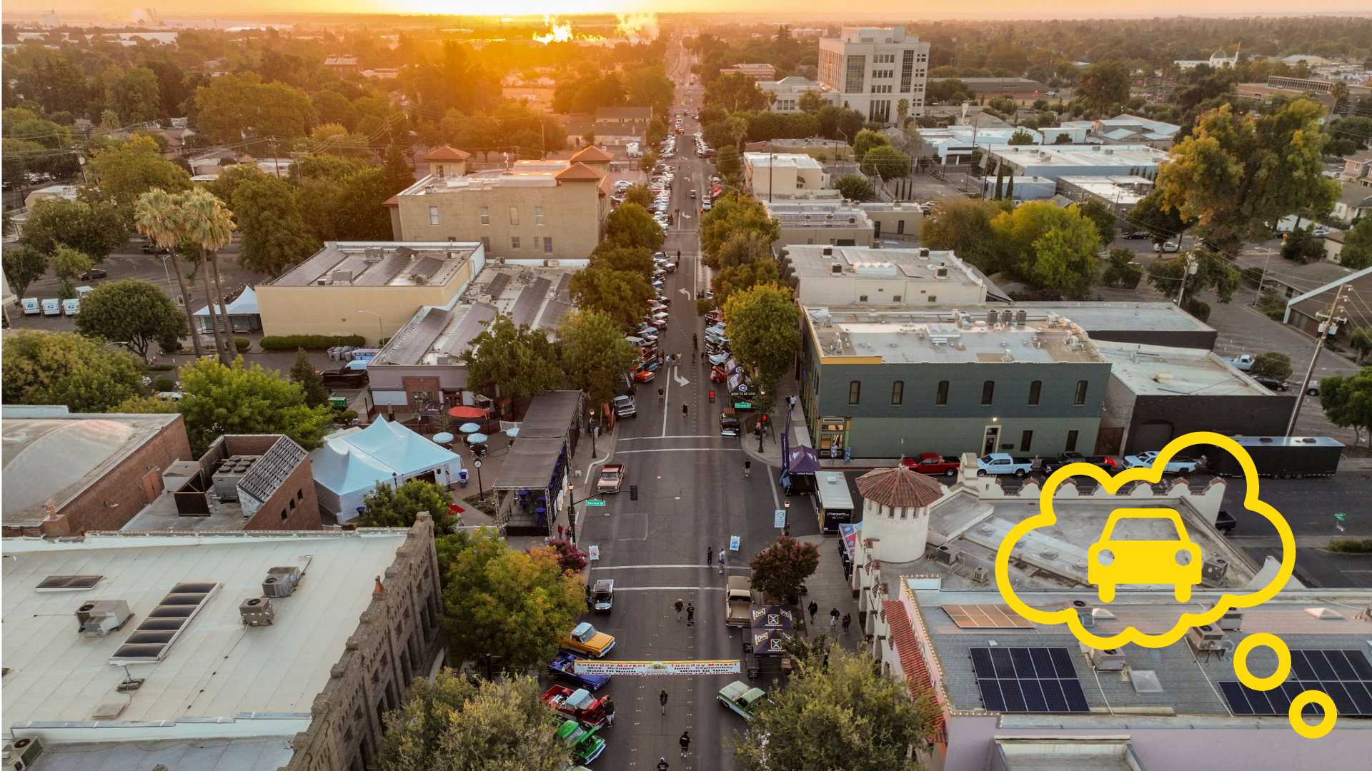 Aerial photograph of a city street at sunset showing buildings, trees, and parked cars lining both sides of the road.