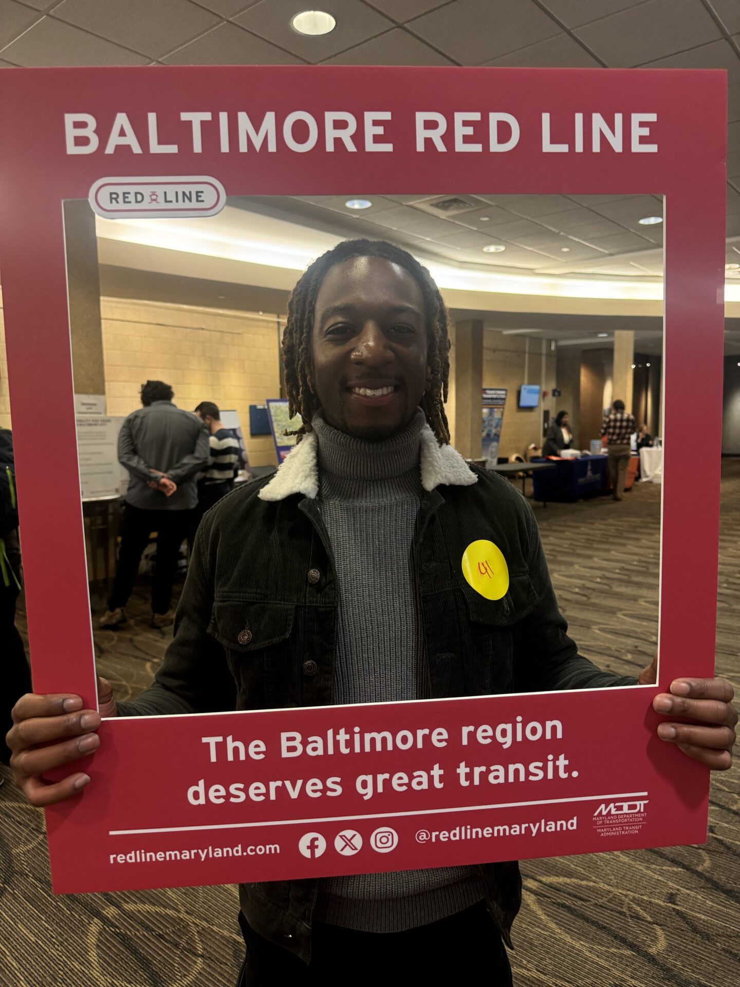 Photograph of a person holding a red promotional frame for Baltimore Red Line transit project at an indoor event. Frame includes text 