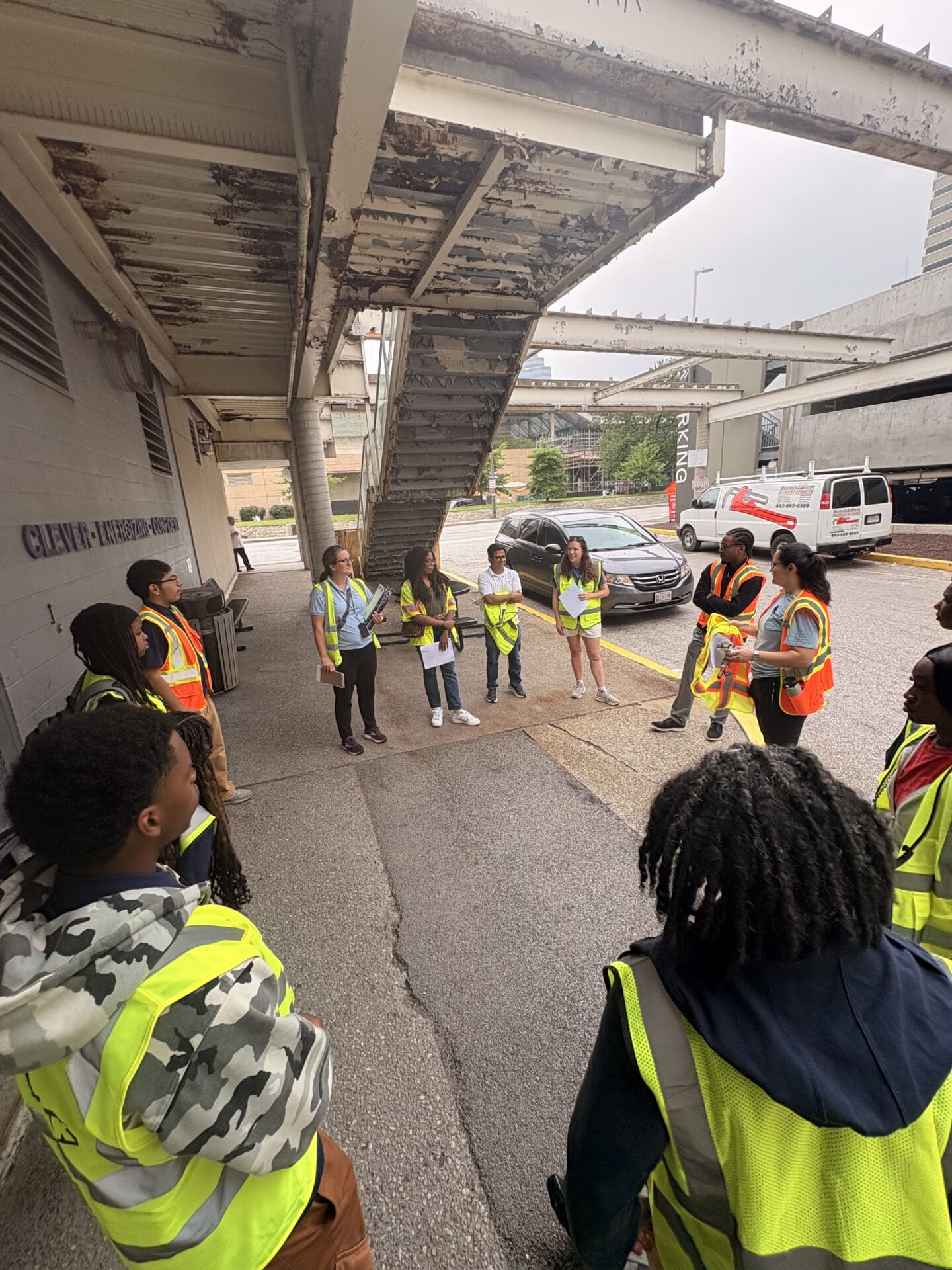 Photograph of a group of people wearing safety vests standing in a circle under a weathered staircase in an outdoor setting.