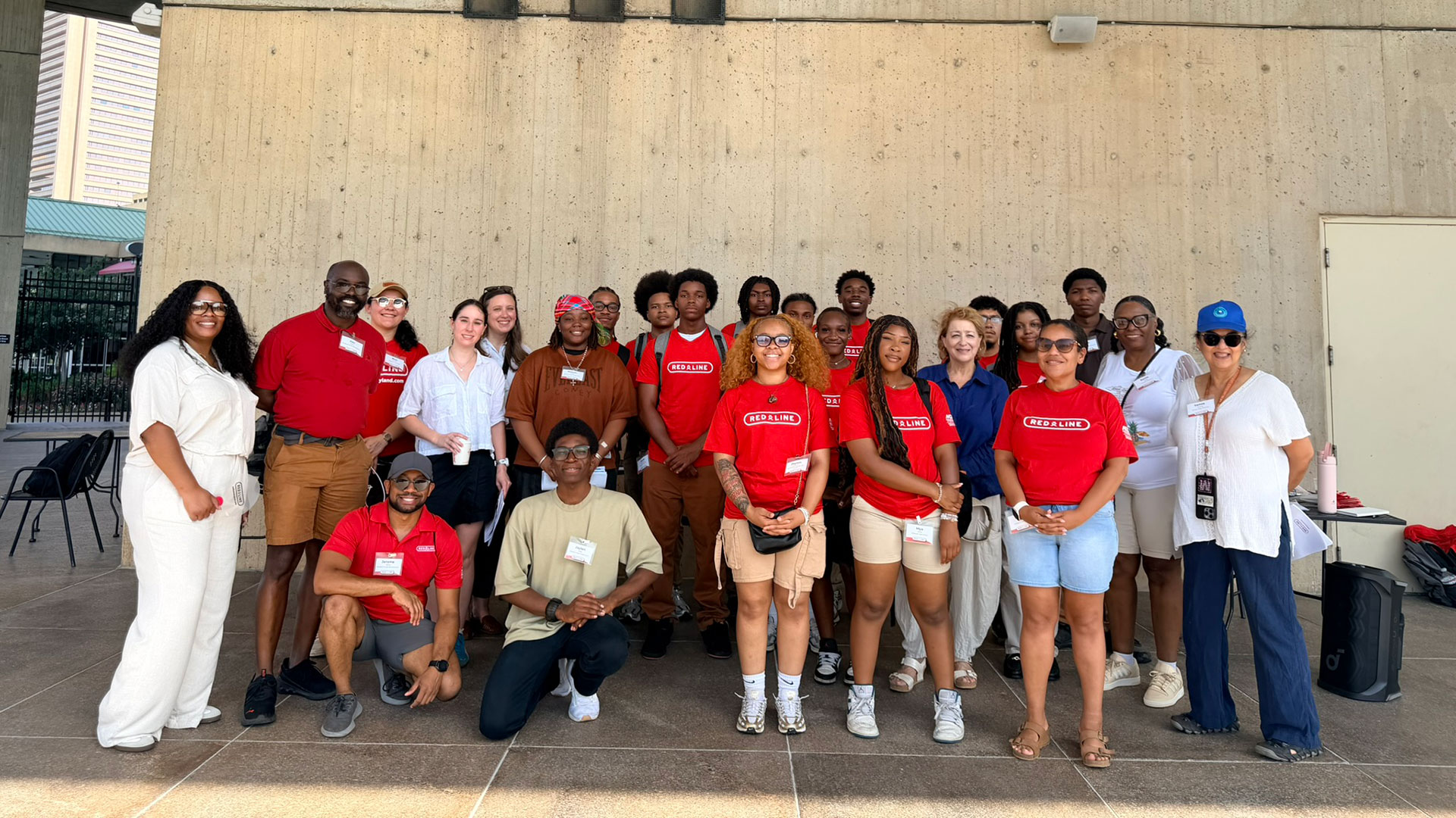 Photograph of a diverse group of 22 people posing together in front of a concrete wall, some wearing red shirts with white text and others in casual attire.