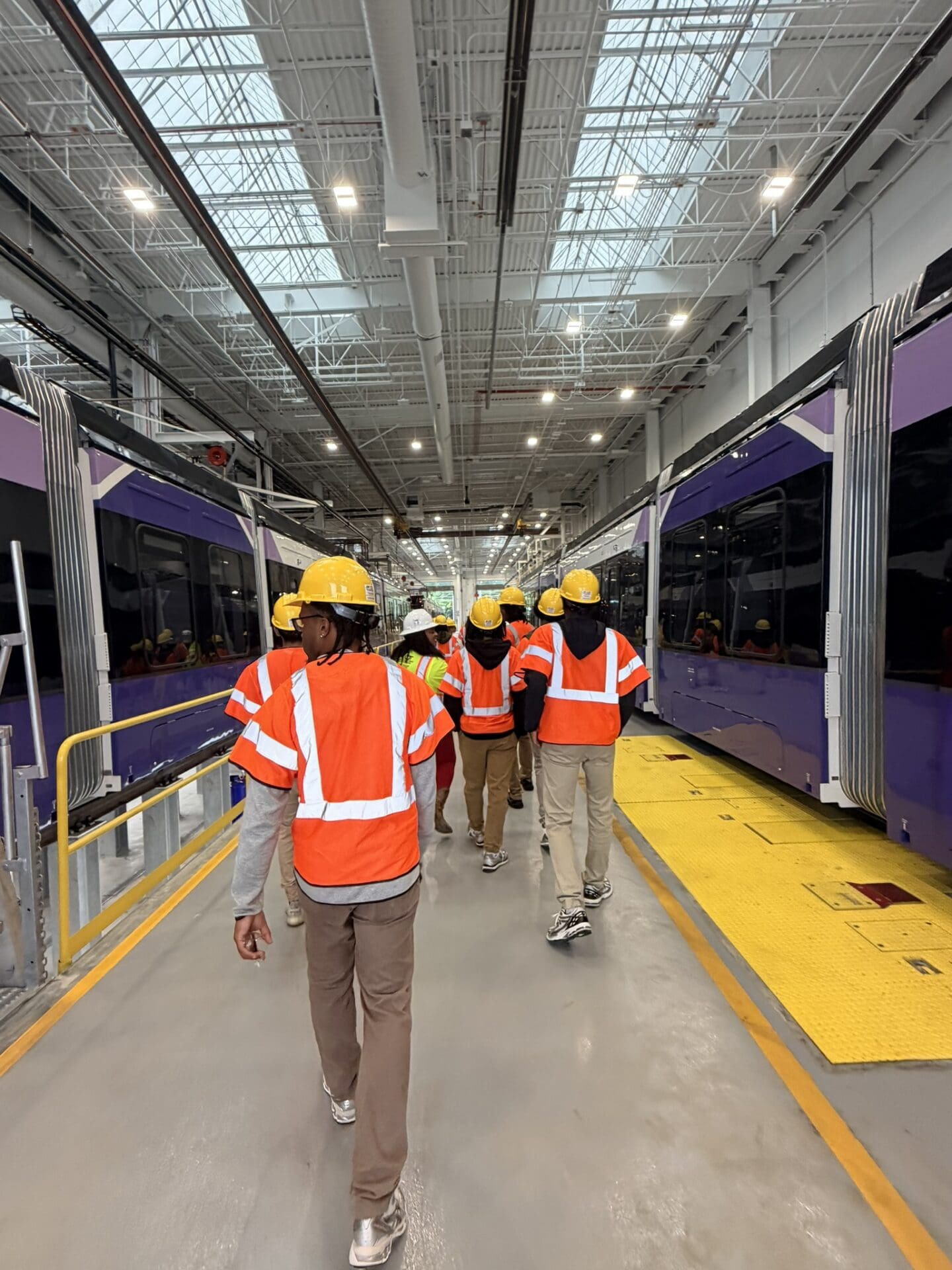 Photograph showing a group of people wearing orange safety vests and yellow hard hats walking between two purple trains inside a large, well-lit maintenance facility. The facility features high ceilings with skylights, industrial lighting, and safety markings on the floor.