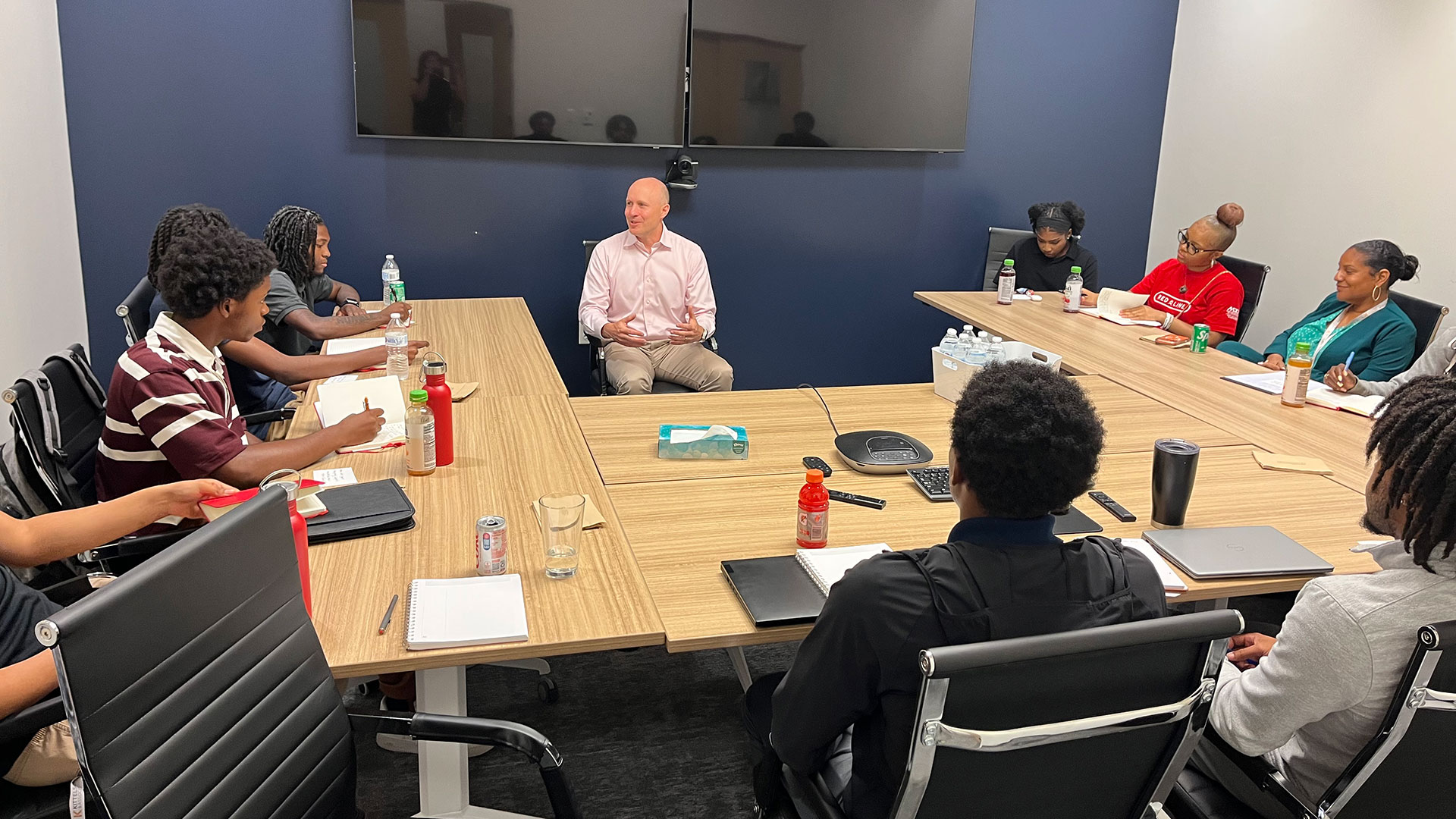 Photograph of a business meeting with nine people seated around a conference table, on a man in a light pink shirt is speaking at the front.