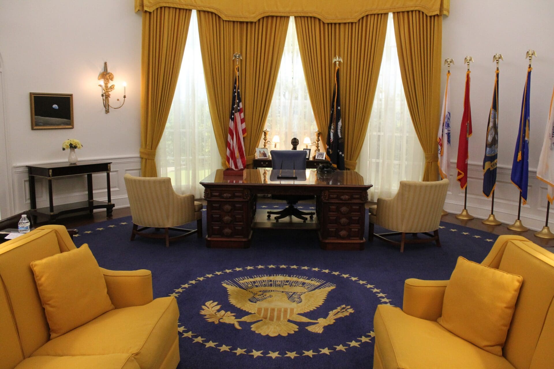 Photograph of the Oval Office with a large wooden desk centered between two beige chairs, with two yellow armchairs in the foreground. The room includes gold curtains, an American flag, military flags, a blue carpet with a presidential seal, and classic wall sconces.