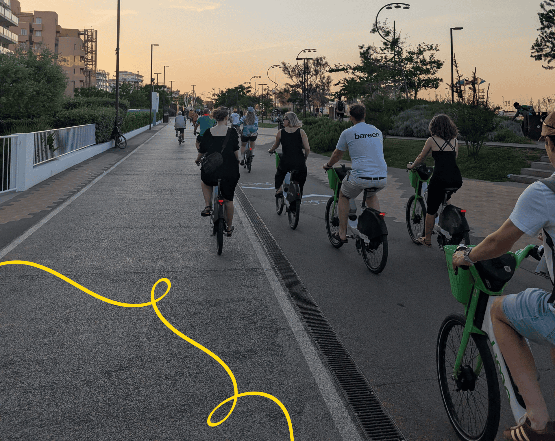 Photograph of a group of people riding bicycles on a paved path during sunset, with buildings on the left and trees on the right.