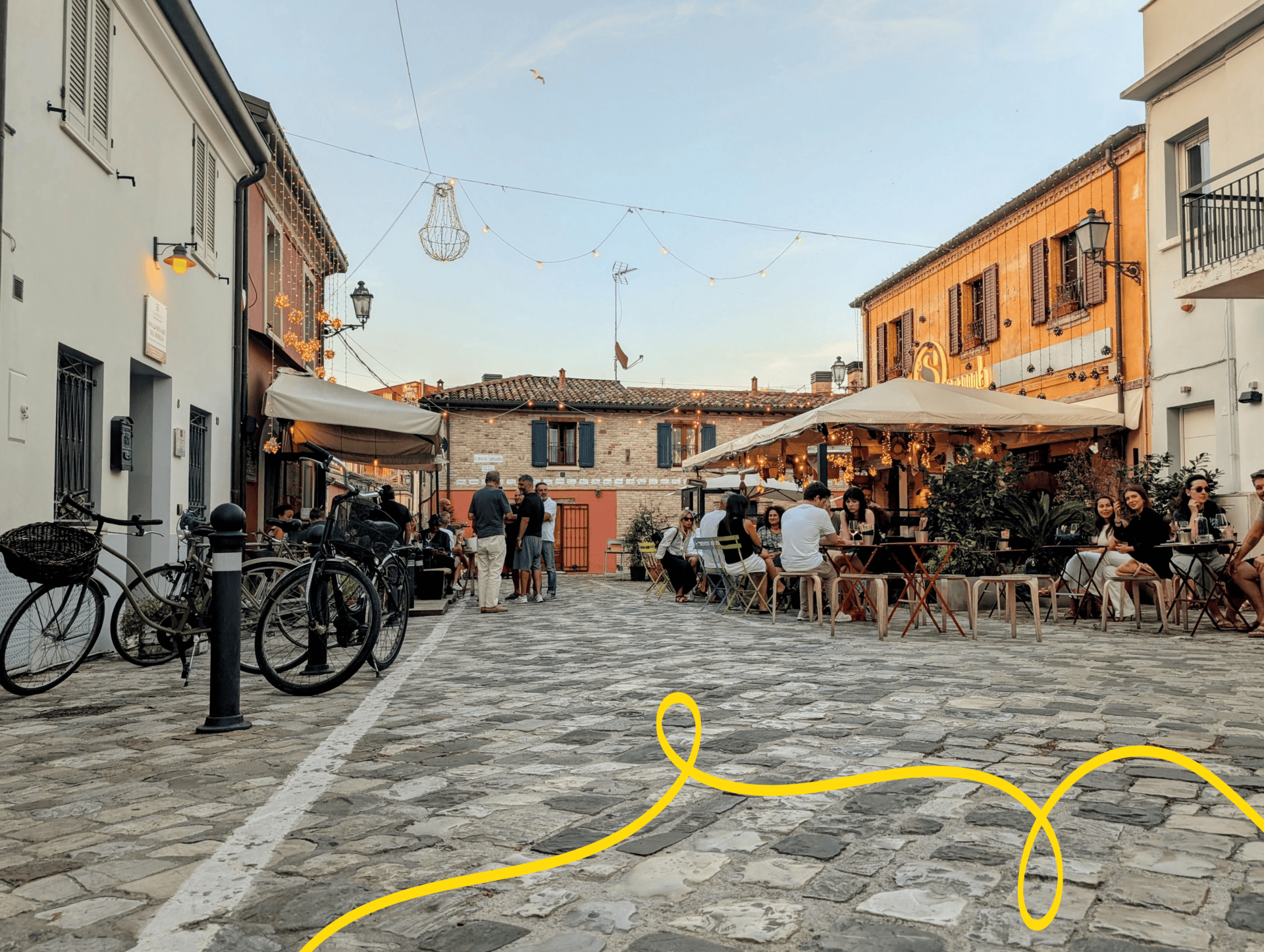 Photograph of a cobblestone street scene featuring outdoor café seating with people dining and socializing under large umbrellas.