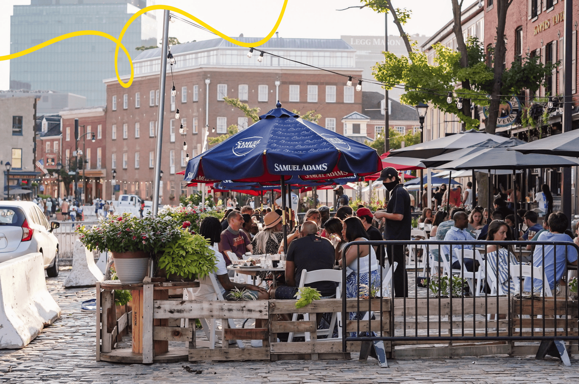 Photograph of a lively street-side dining area with people seated under a blue Samuel Adams umbrella and adjacent patio umbrellas. The scene features cobblestone pavement, potted plants, and historic brick buildings.