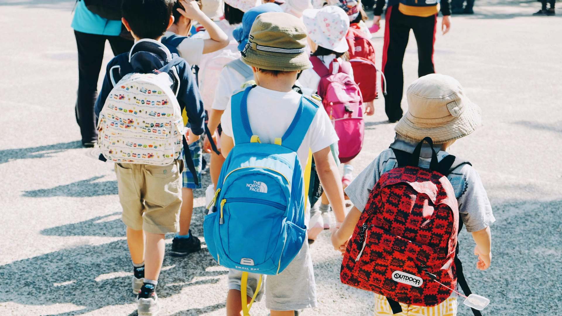 Photograph of young children walking outdoors, each wearing colorful backpacks and sun hats. Bright sunlight casts shadows on pavement.