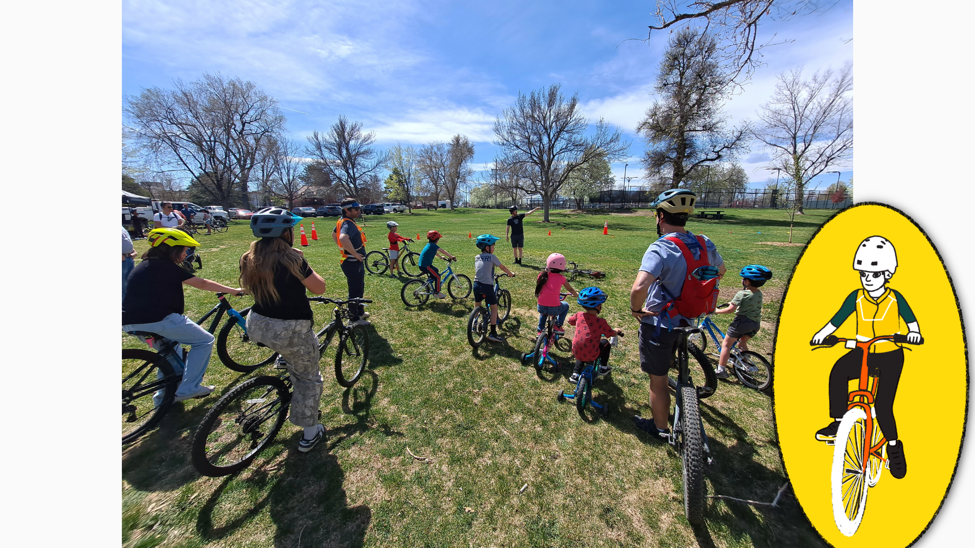 A photo of several children on bikes lined up on a grassy field. Two adult chaperones supervise them in an activity zone demarcated by traffic cones.